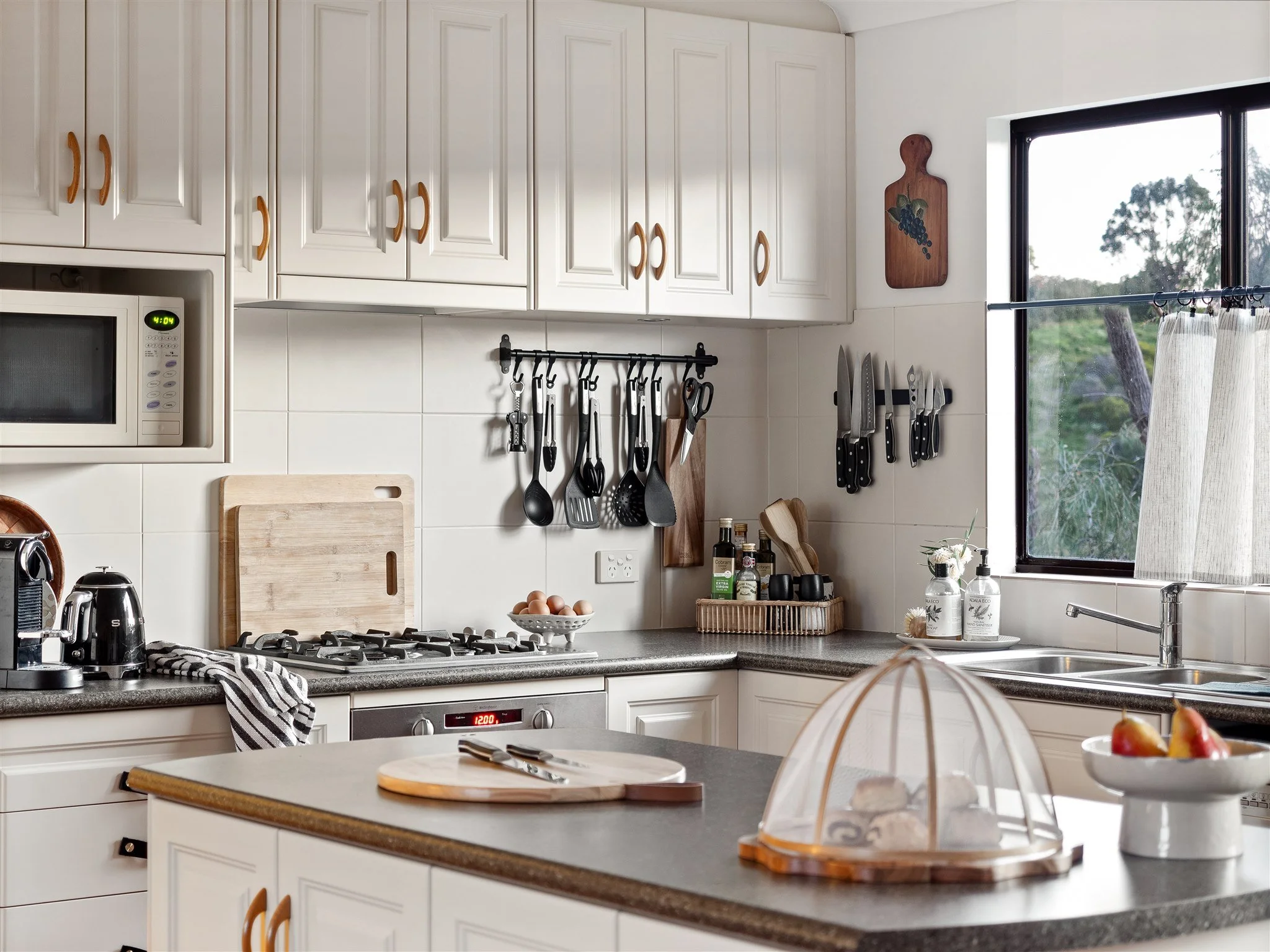 A kitchen with white cabinets, a window with a view of trees, and various kitchen utensils hanging on the wall.
