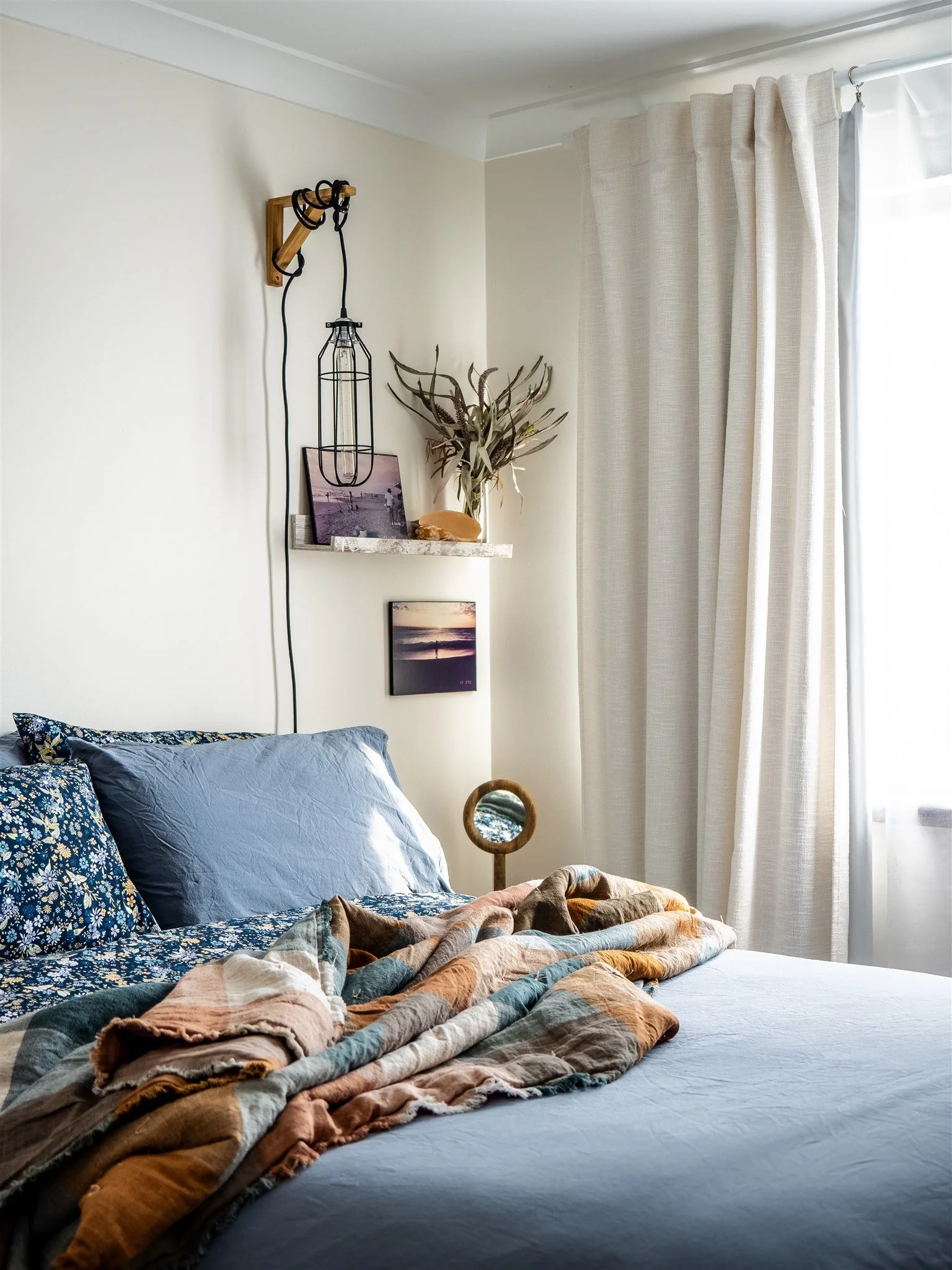A neatly made bed with a blue floral pillow, a solid blue pillow, and a multicolored quilt. There is a small round mirror on a wooden stand beside the bed. The wall above the bed has a floating shelf with a framed photo, a dried plant arrangement, an