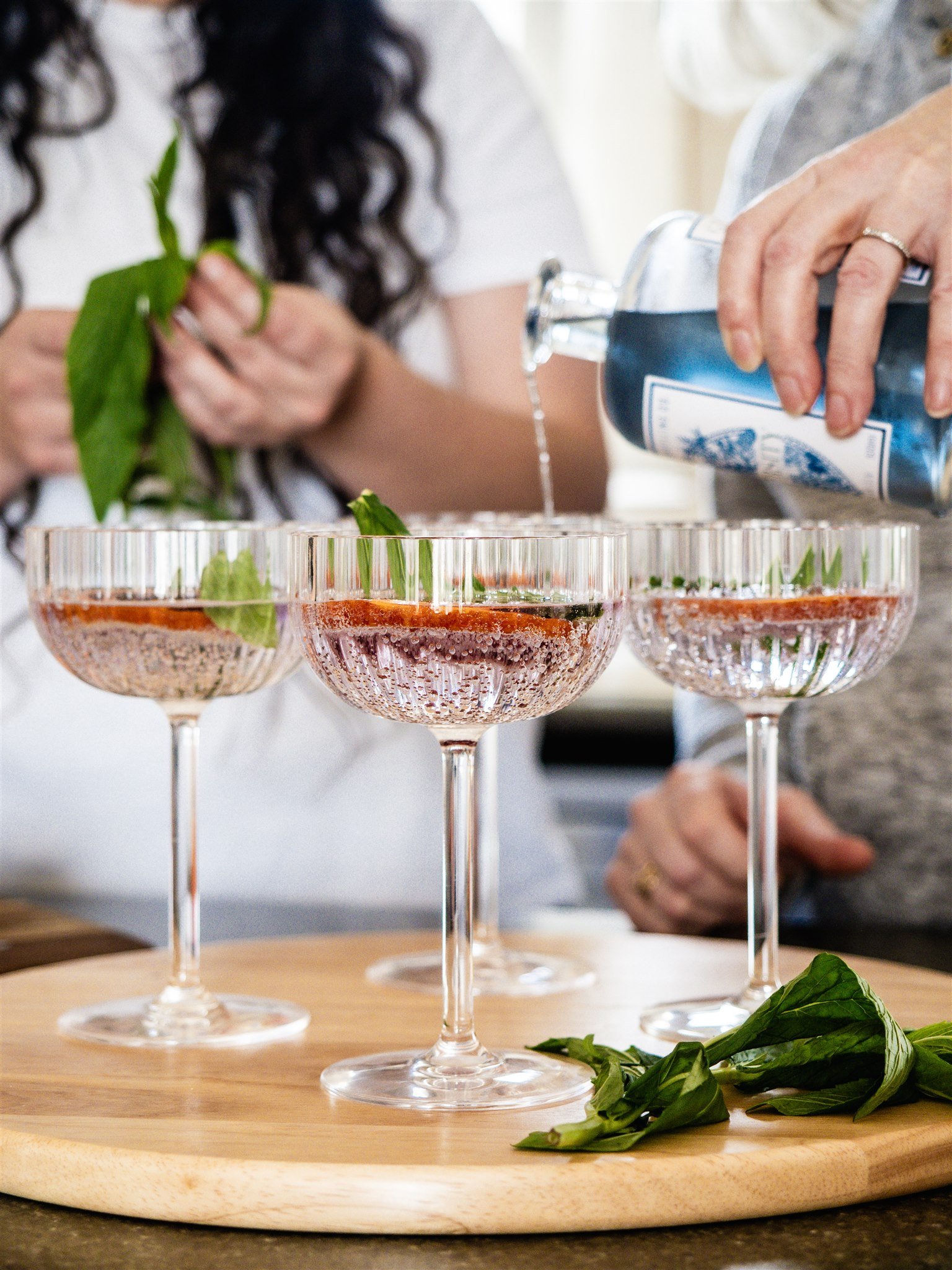Close-up of three pink cocktails topped with basil leaves in vintage coupe glasses, with a person pouring blue liquid into one of the drinks, on a wooden tray on a dark counter, with some basil leaves scattered around.