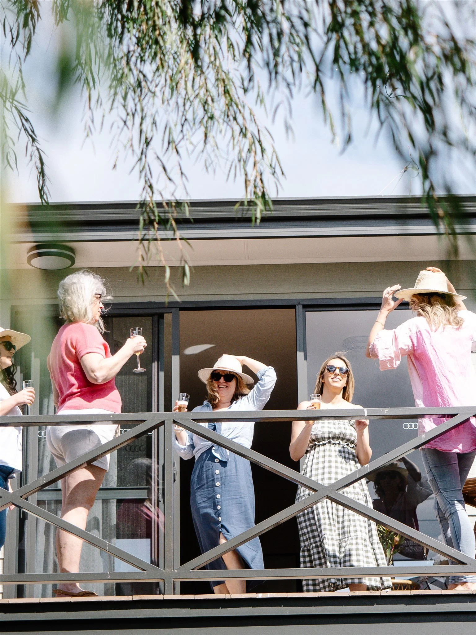 Group of women enjoying drinks on a balcony during daytime, with some wearing hats and sunglasses, and a tree overhead.
