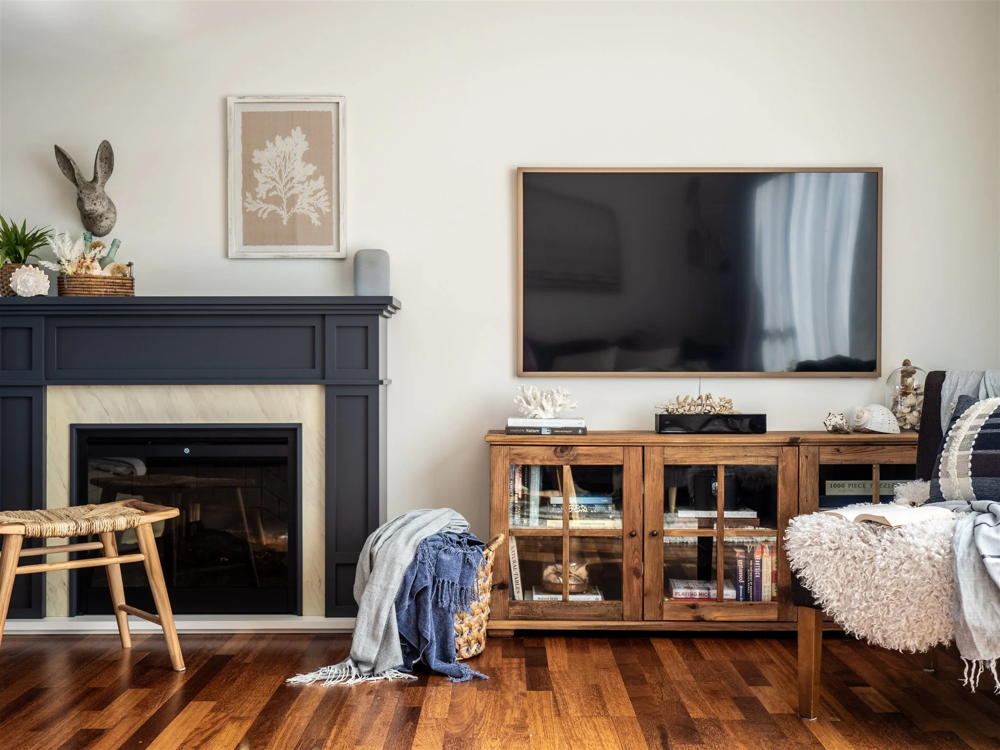 Living room with a fireplace, a mounted decorative rabbit sculpture, framed artwork with a tree, a flat-screen TV, a wooden bookshelf with books, and various decorative items and clothing on furniture and chairs.