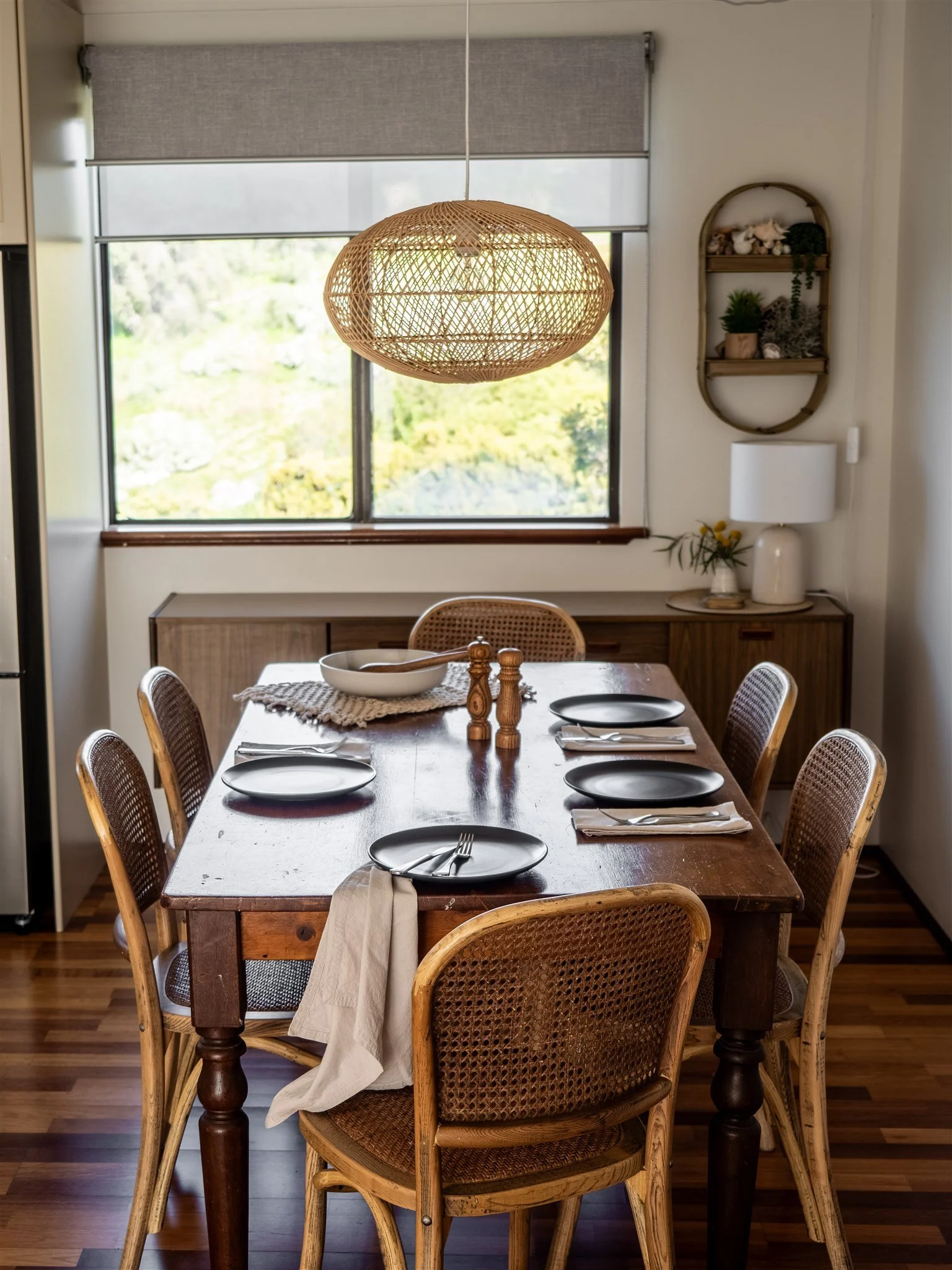 A wooden dining table set for six people with black plates, silverware, and napkins, in a room with a window, a wooden sideboard, a wicker pendant light, and a wall shelf with plants and decorative items.