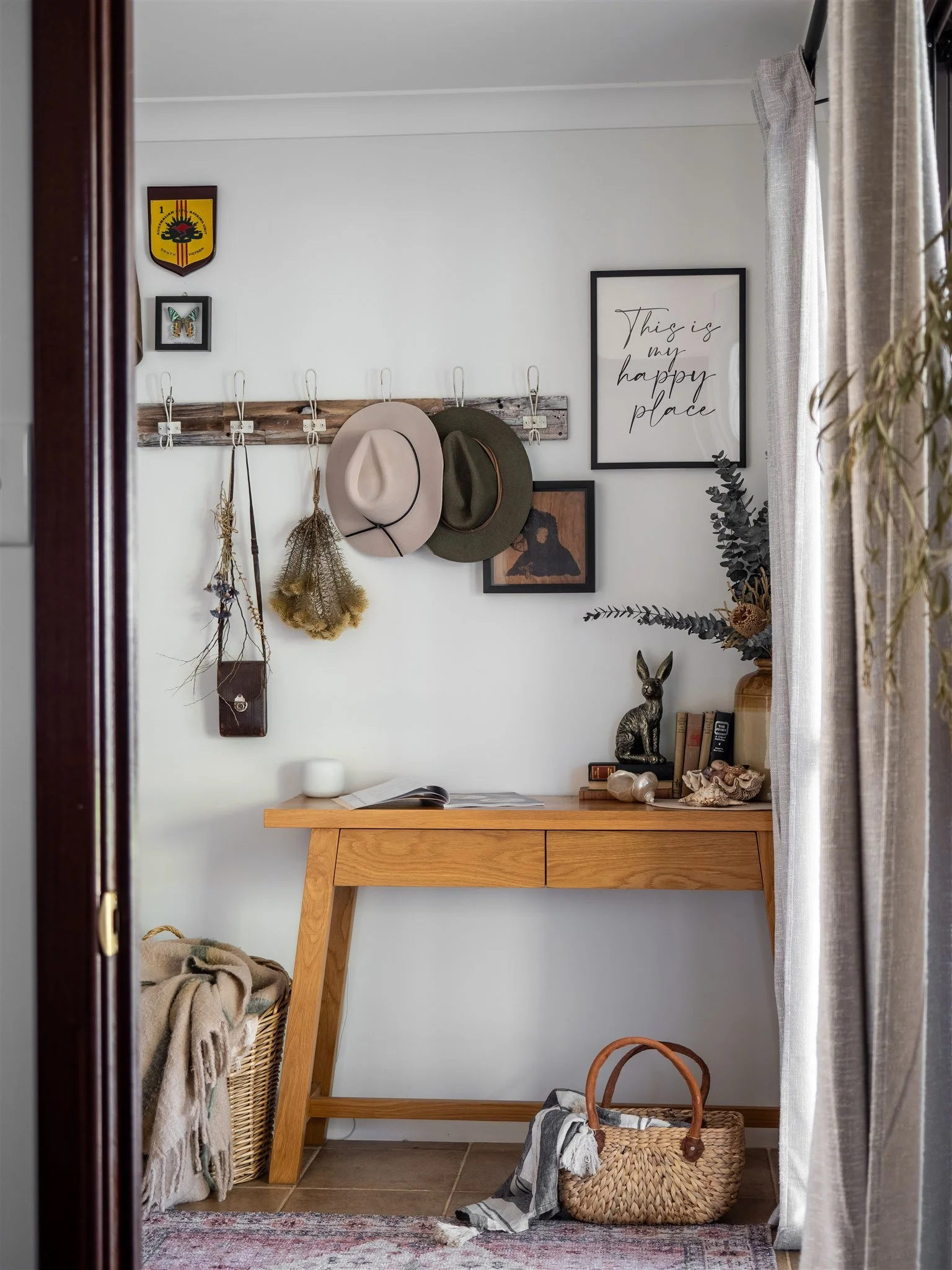 A cozy entryway with a wooden plank coat rack holding two hats, a framed quote that says 'This is my happy place,' and various decorations including dried plants, artwork, and a rabbit figurine. There's a basket with blankets and a woven bag on the f