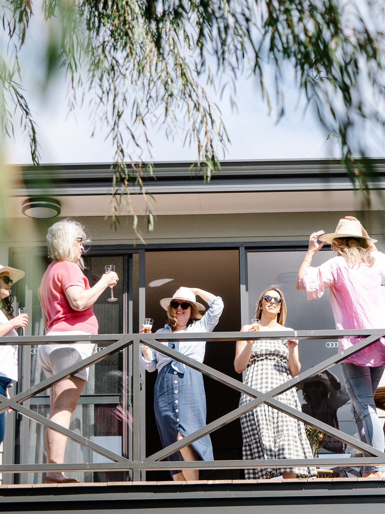 Group of women gathered on a balcony, some holding drinks, wearing summer clothing and hats, smiling and talking, with tree branches overhead.