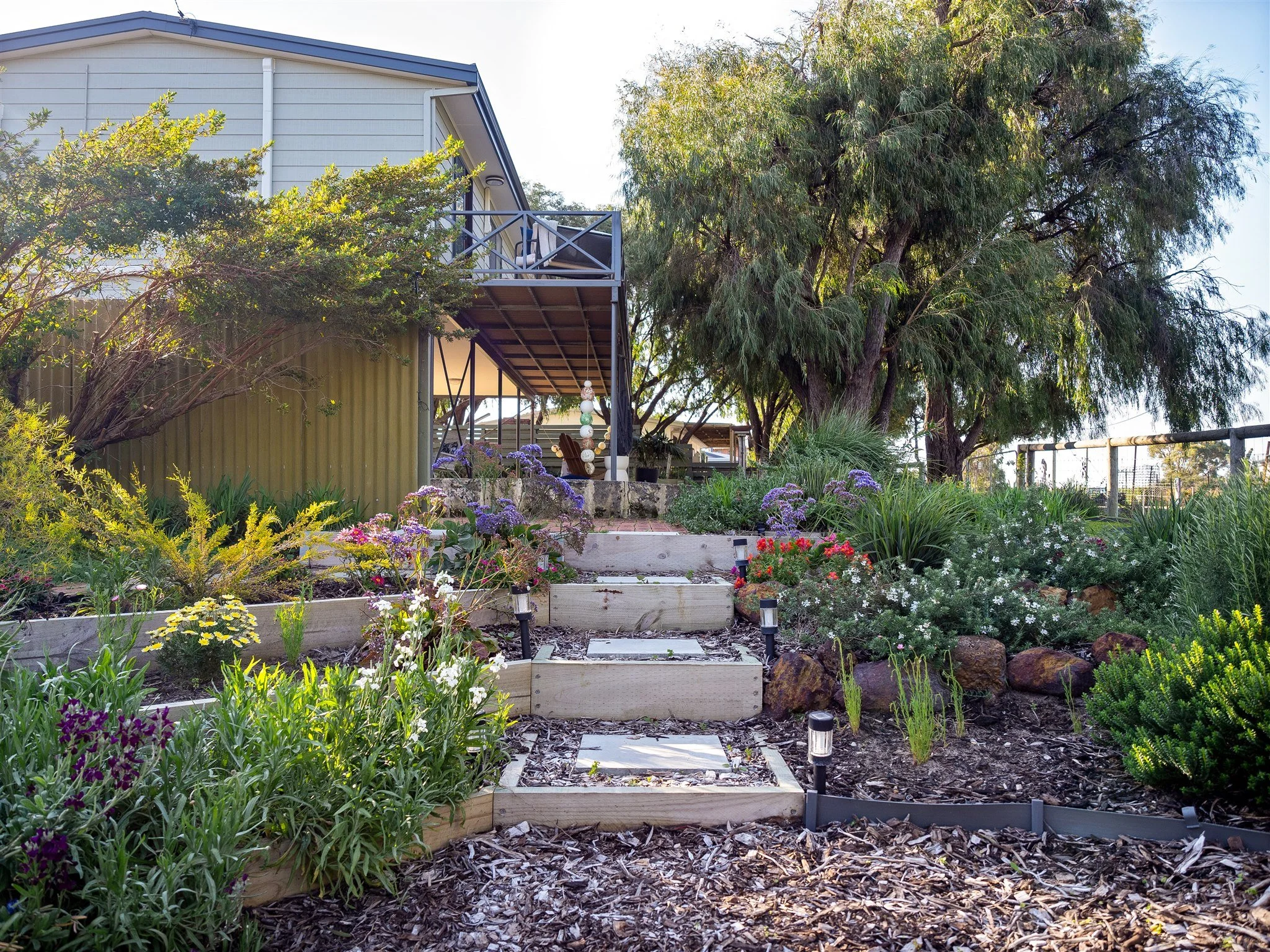 A lush garden with flowers, shrubs, and trees, featuring a raised garden bed, brick pathway, and a two-story house with a porch and railing in the background.