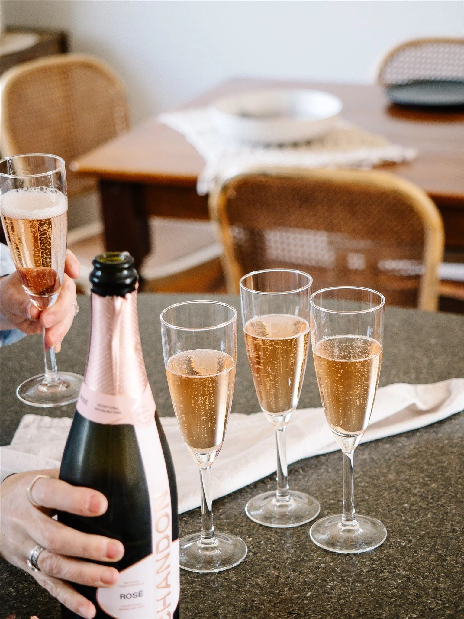 Person holding a bottle of rosé sparkling wine near four filled champagne flutes on a table.