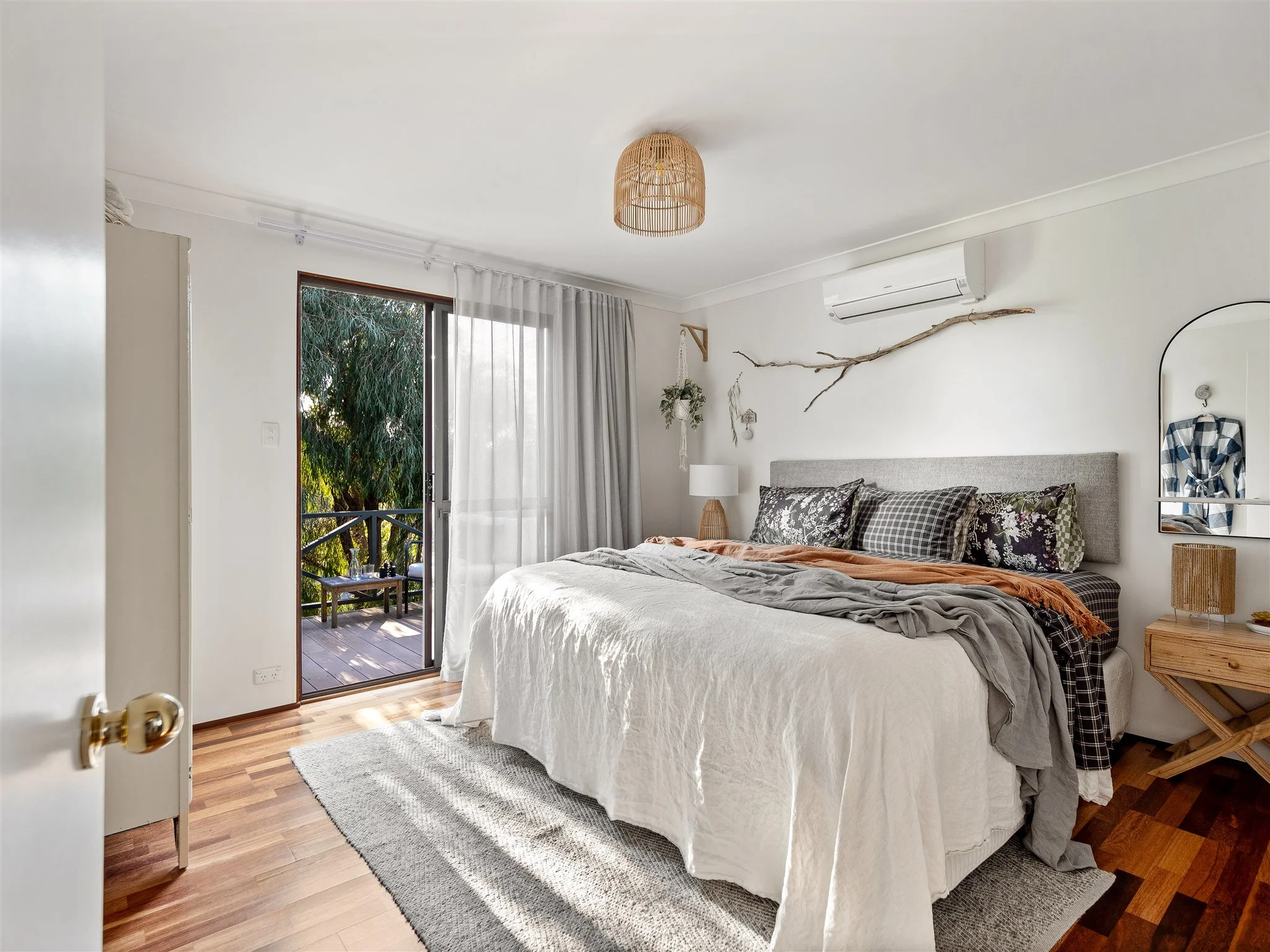 A cozy bedroom with a large bed covered in white and patterned bedding. There are curtains and a sliding glass door leading to a balcony with outdoor seating. Natural light illuminates the room, which has wood flooring, a small bedside table, and a w