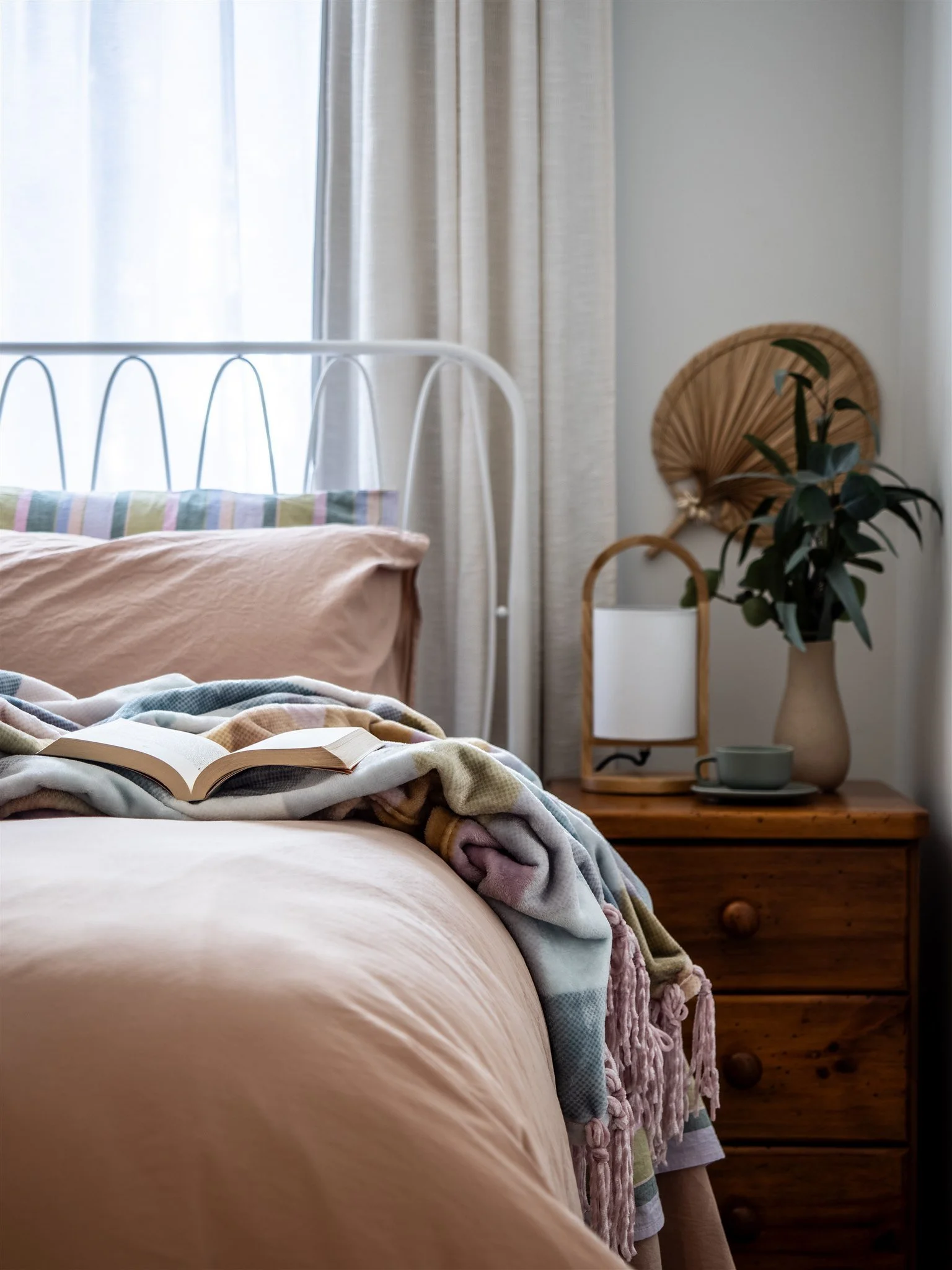 A cozy bedroom scene featuring a bed with a peach-colored duvet and a colorful striped pillow. An open book rests on a blanket at the foot of the bed. In the background, a wooden nightstand with a large vase holding a leafy green plant, a white decor