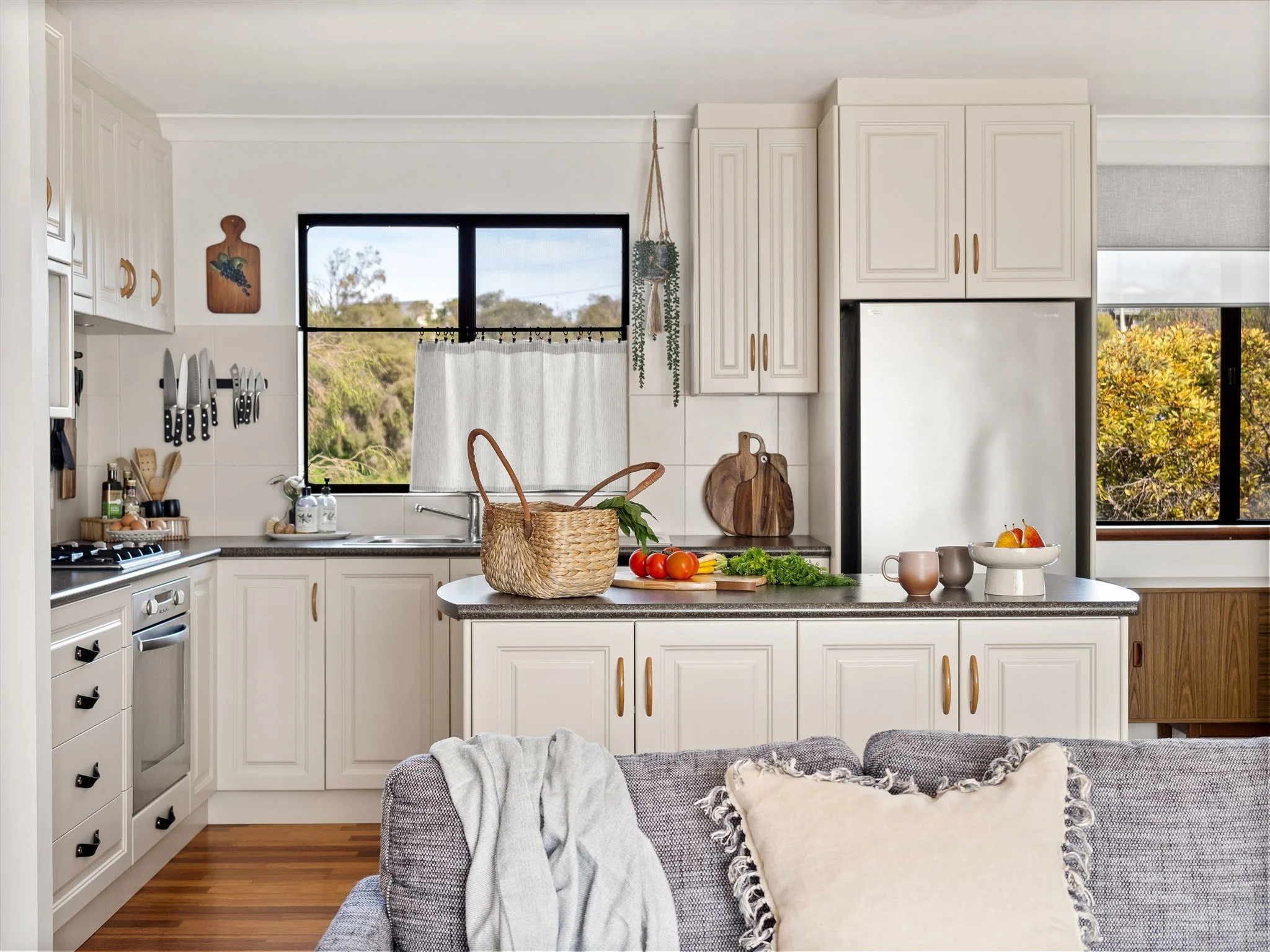 A modern kitchen with white cabinets, a window with a view of trees, and a gray sofa in the living area.