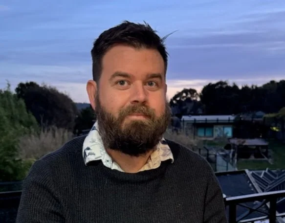 A man with dark hair and a beard, wearing a black sweater over a collared shirt, stands outdoors during sunset with a background of trees, buildings, and a partly cloudy sky.