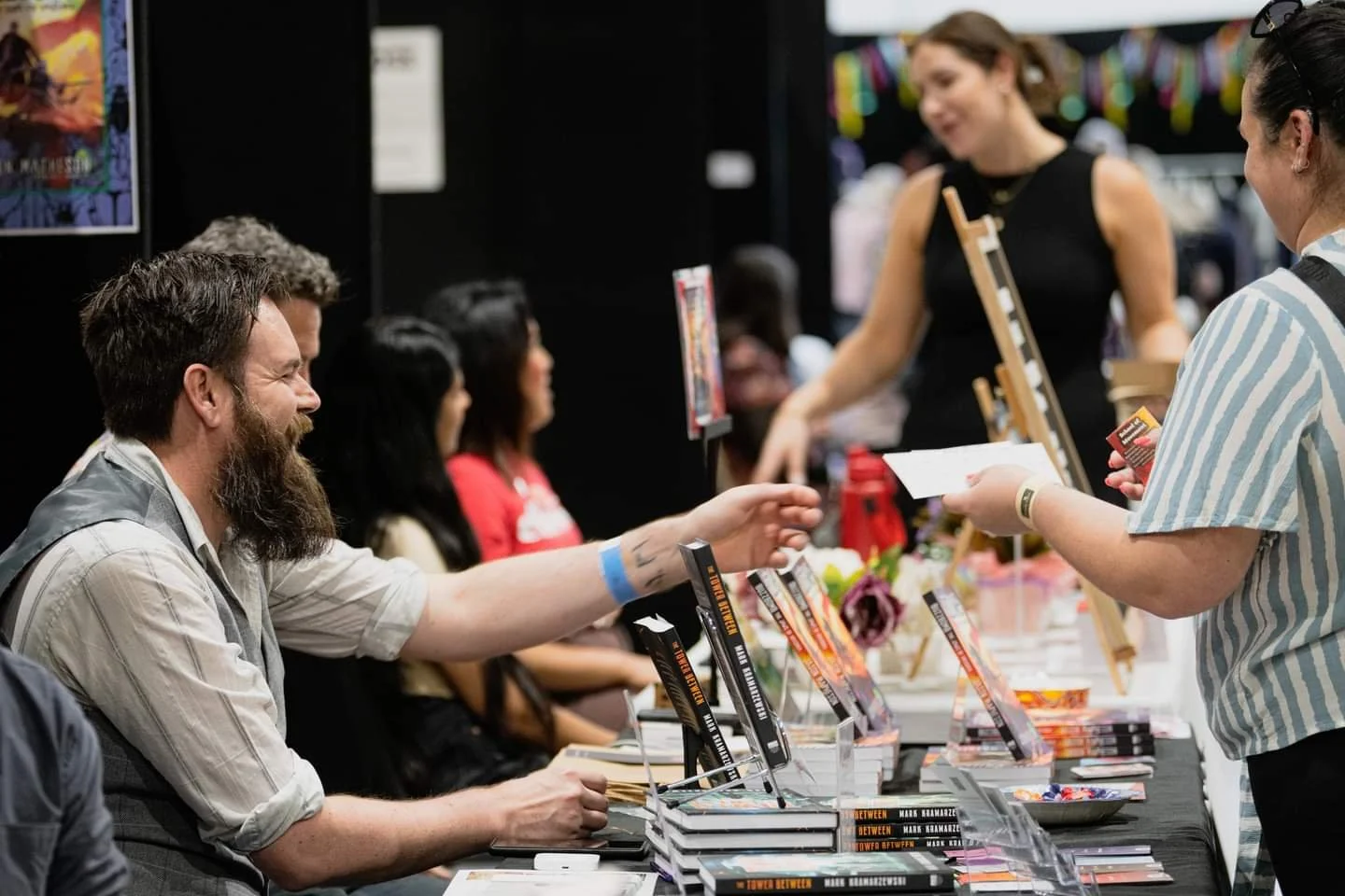 A scene at a book signing event with a man with a beard and tattoos, sitting at a table and signing a book for a woman, while other people sit at the table and a woman stands and interacts with attendees.