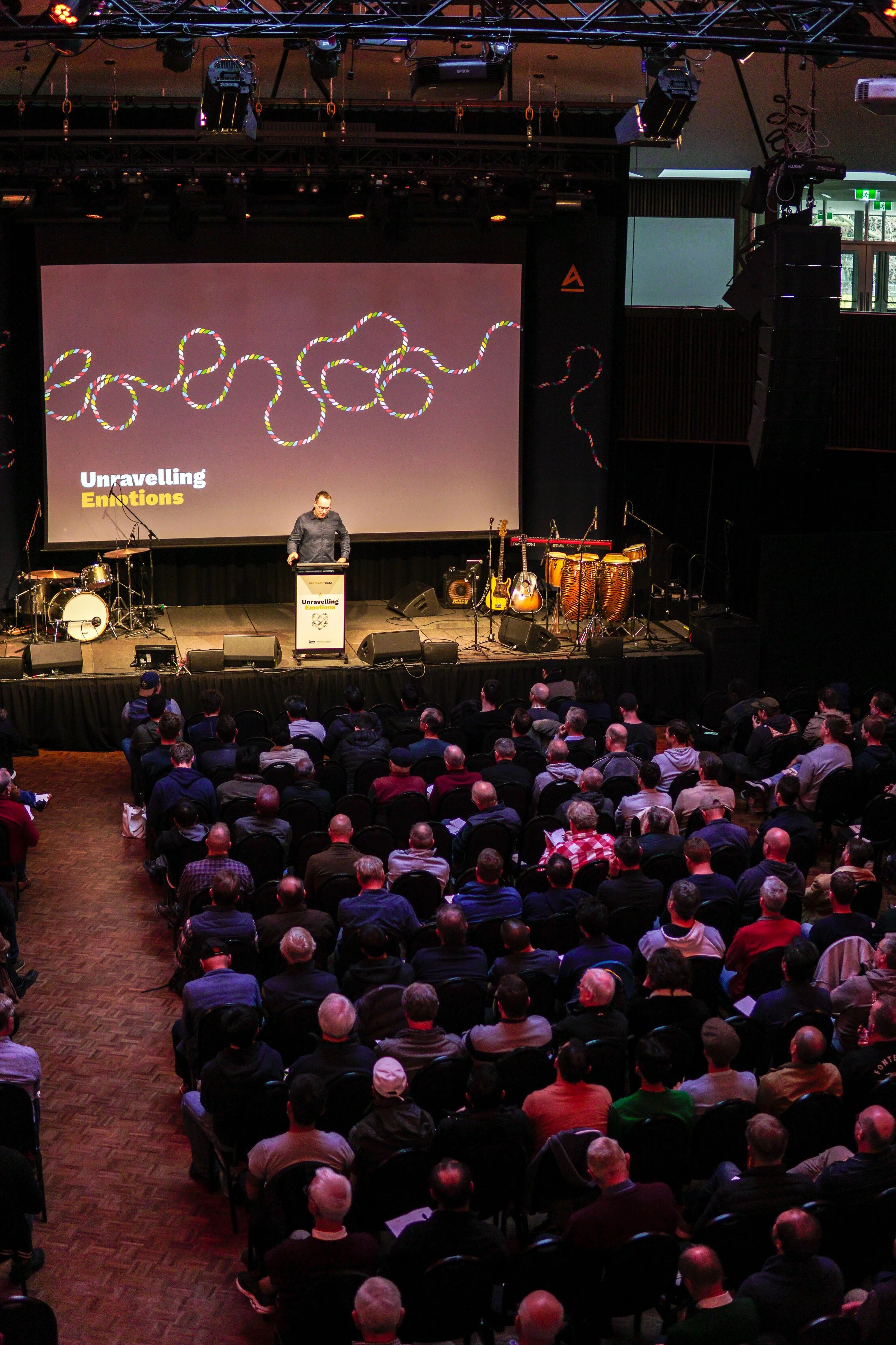 An indoor stage with a man standing behind a podium during a presentation. The backdrop shows a graphic of multicolored intertwined ropes and the words 'Unravelling Emotions.' The stage is set with musical instruments, including drums, guitars, and congas. Audience members are seated facing the stage.