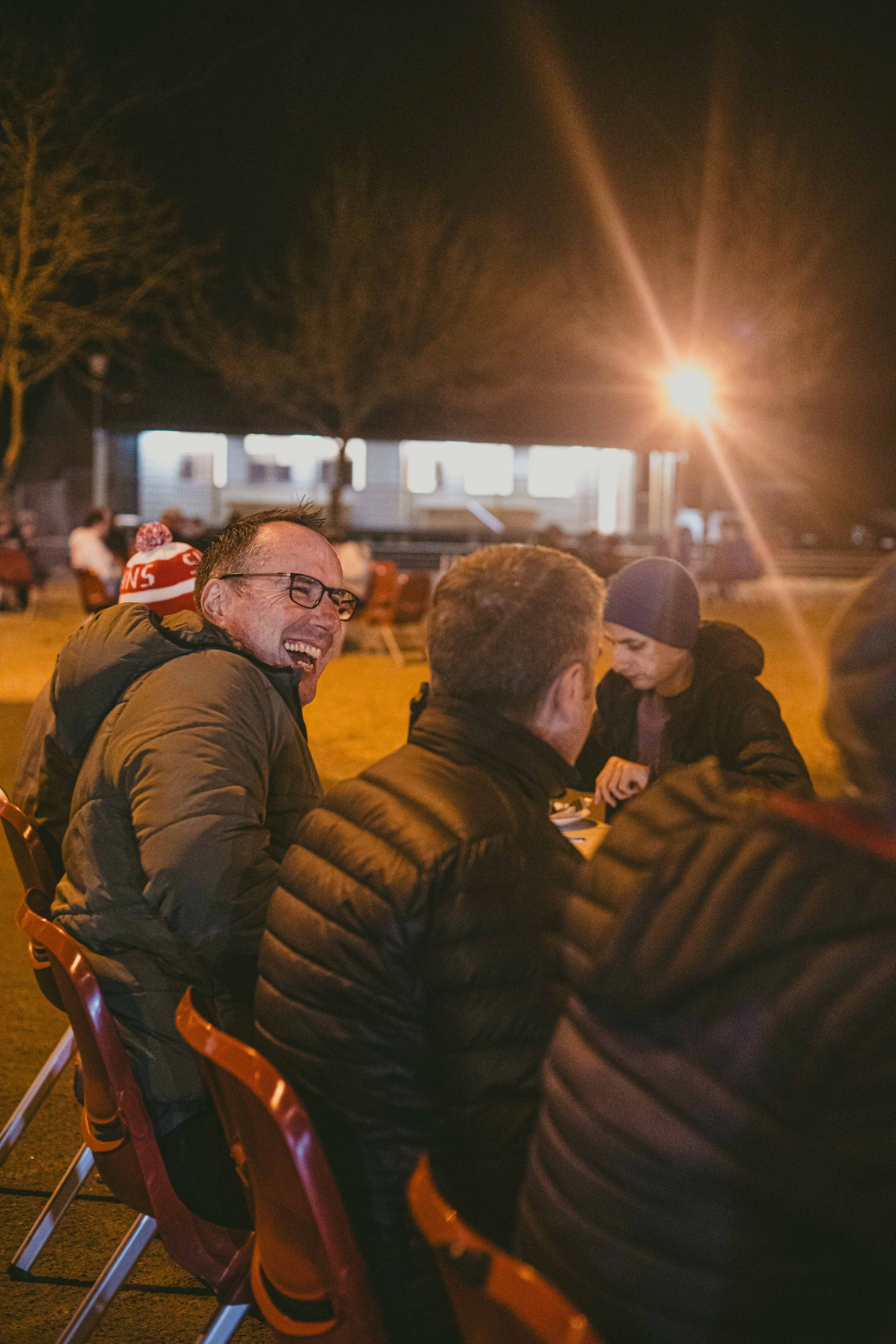 People sitting at outdoor table at night, illuminated by a bright outdoor light, laughing and talking.