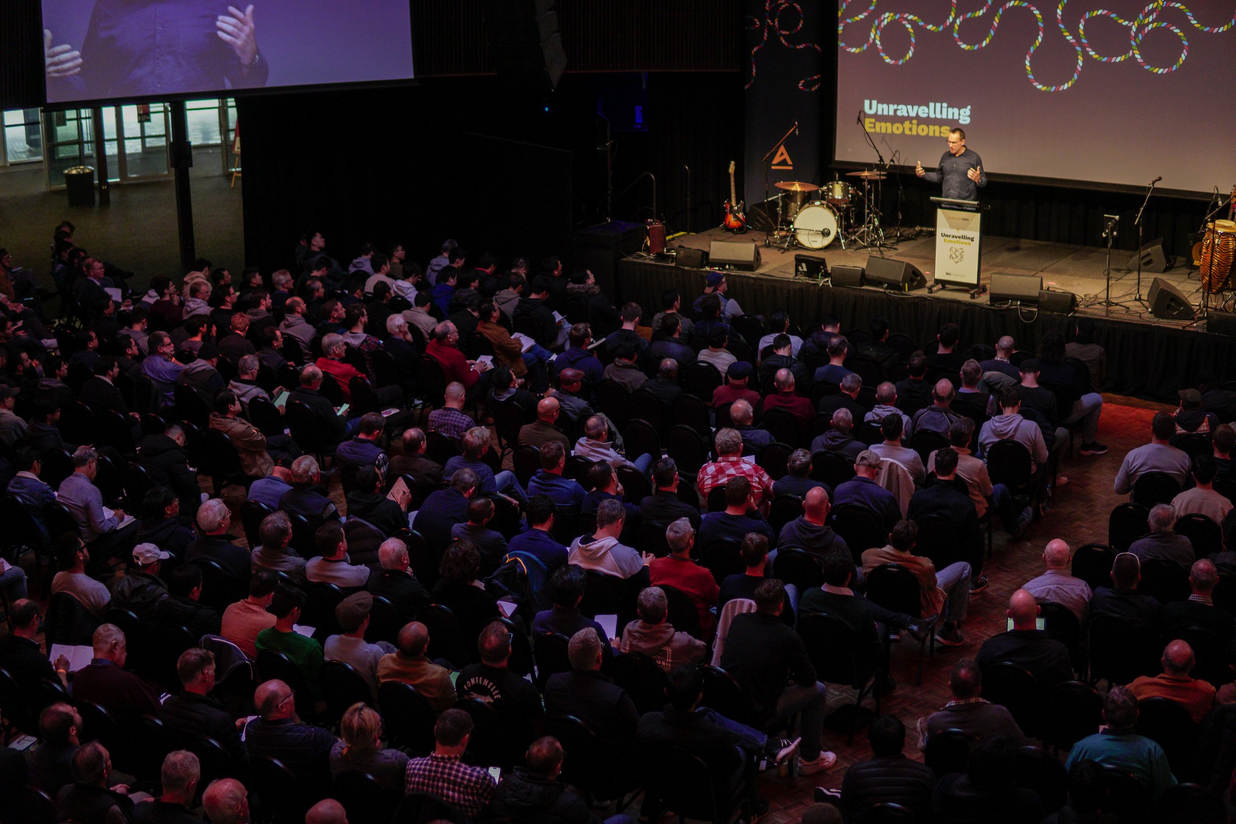 A large indoor auditorium filled with people seated and listening to a speaker on stage. The stage has musical instruments and a large screen with colorful graphics and the words "Unravelling Emotions".
