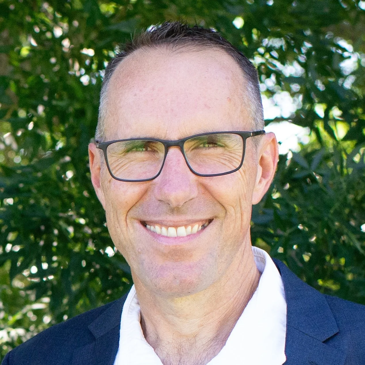 A smiling man with short dark hair, wearing glasses, a white shirt, and a dark blazer, standing outdoors with green foliage in the background.