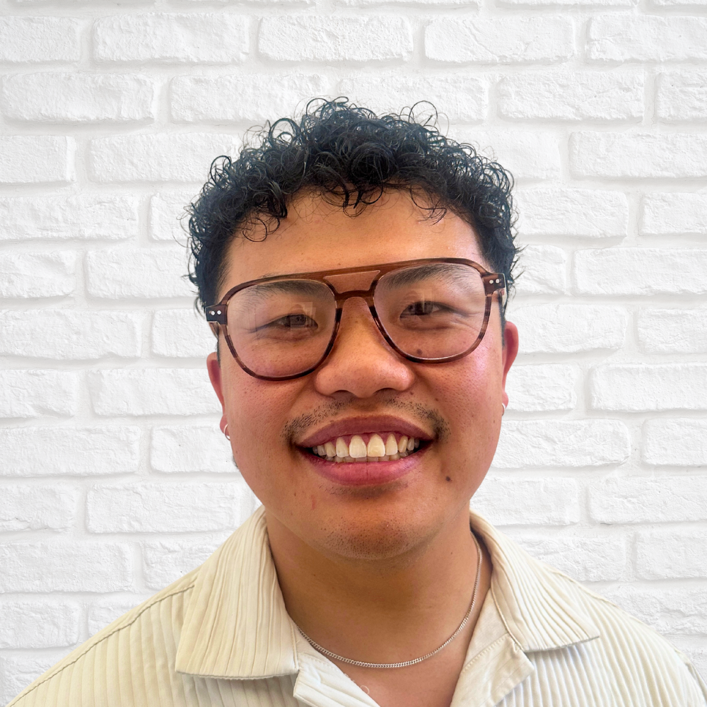 A young man with curly hair, wearing large glasses and a light-colored shirt, smiling in front of a white brick wall.