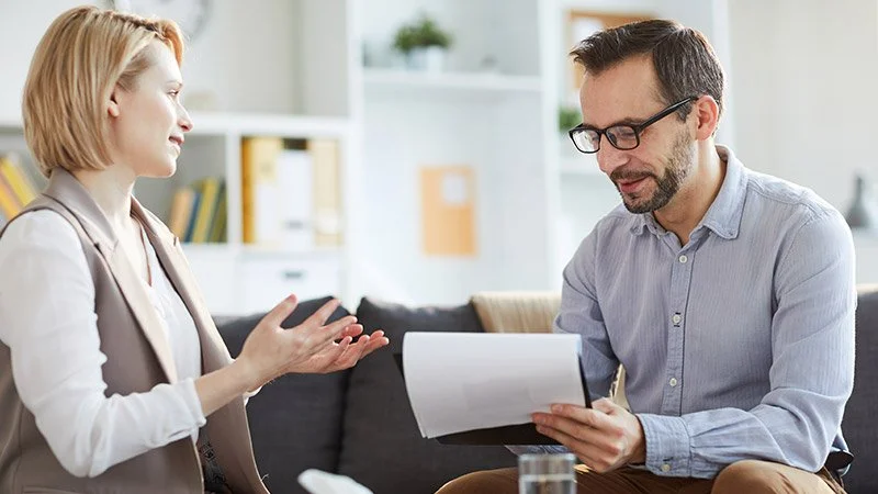 A woman and a man are sitting on a couch in an office, having a conversation. The woman is gesturing with her hands, and the man is holding a notebook, listening attentively.