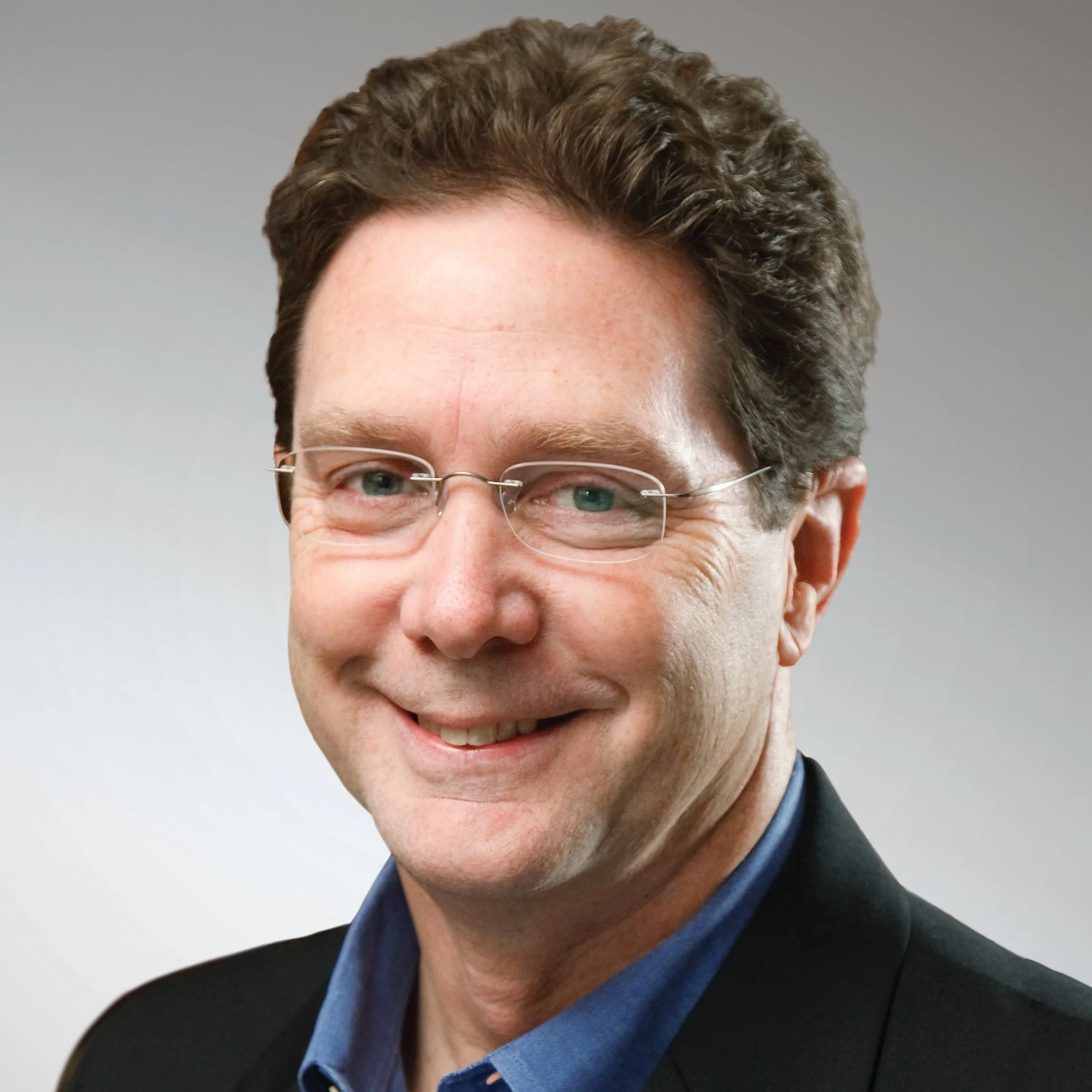 A close-up of a man's face with blue eyes, wearing glasses, a dark suit, and a blue shirt, smiling.