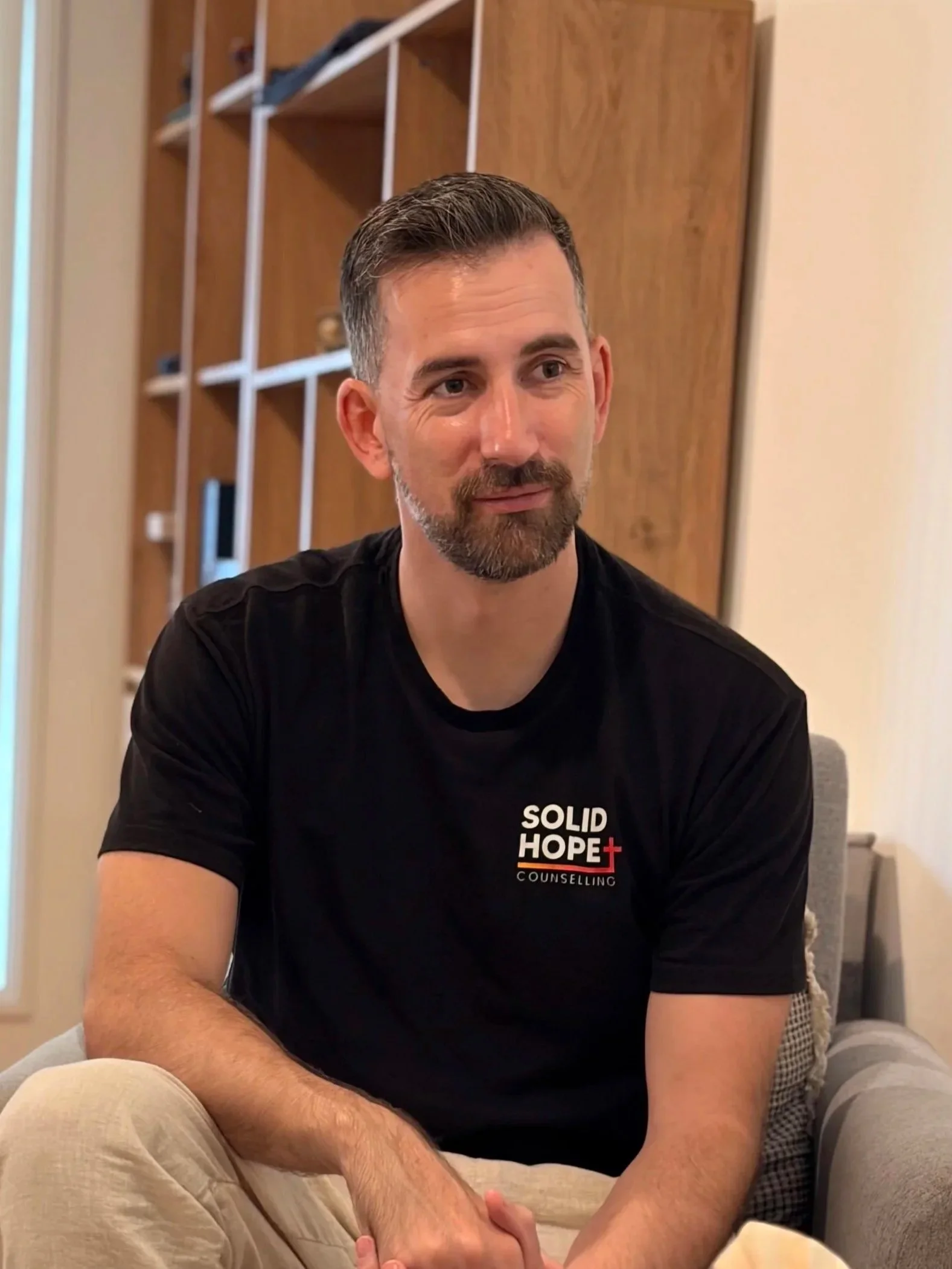 A man with short dark hair and a beard sits on a couch, smiling slightly, wearing a black t-shirt with 'Solid Hope Counseling' printed on it. Behind him is a wooden wall with a white shelving unit and some objects on the shelves.