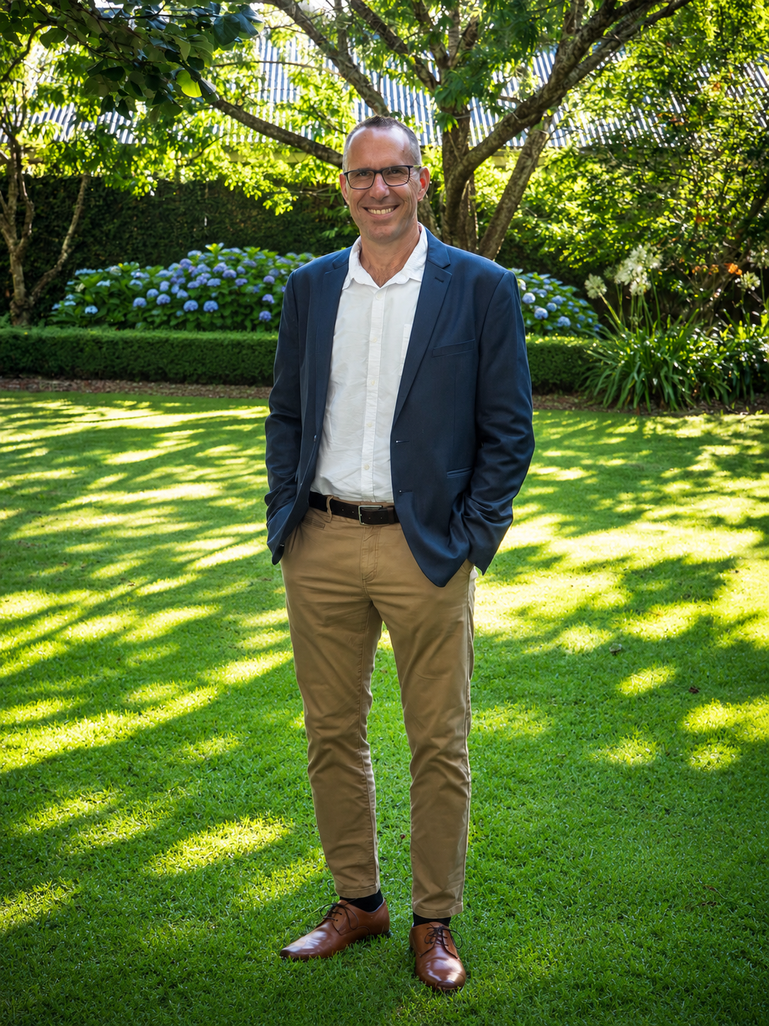 A man in a navy blazer, white shirt, khaki pants, and brown shoes posing outdoors on a sunny day with lush green trees and flowers in the background.
