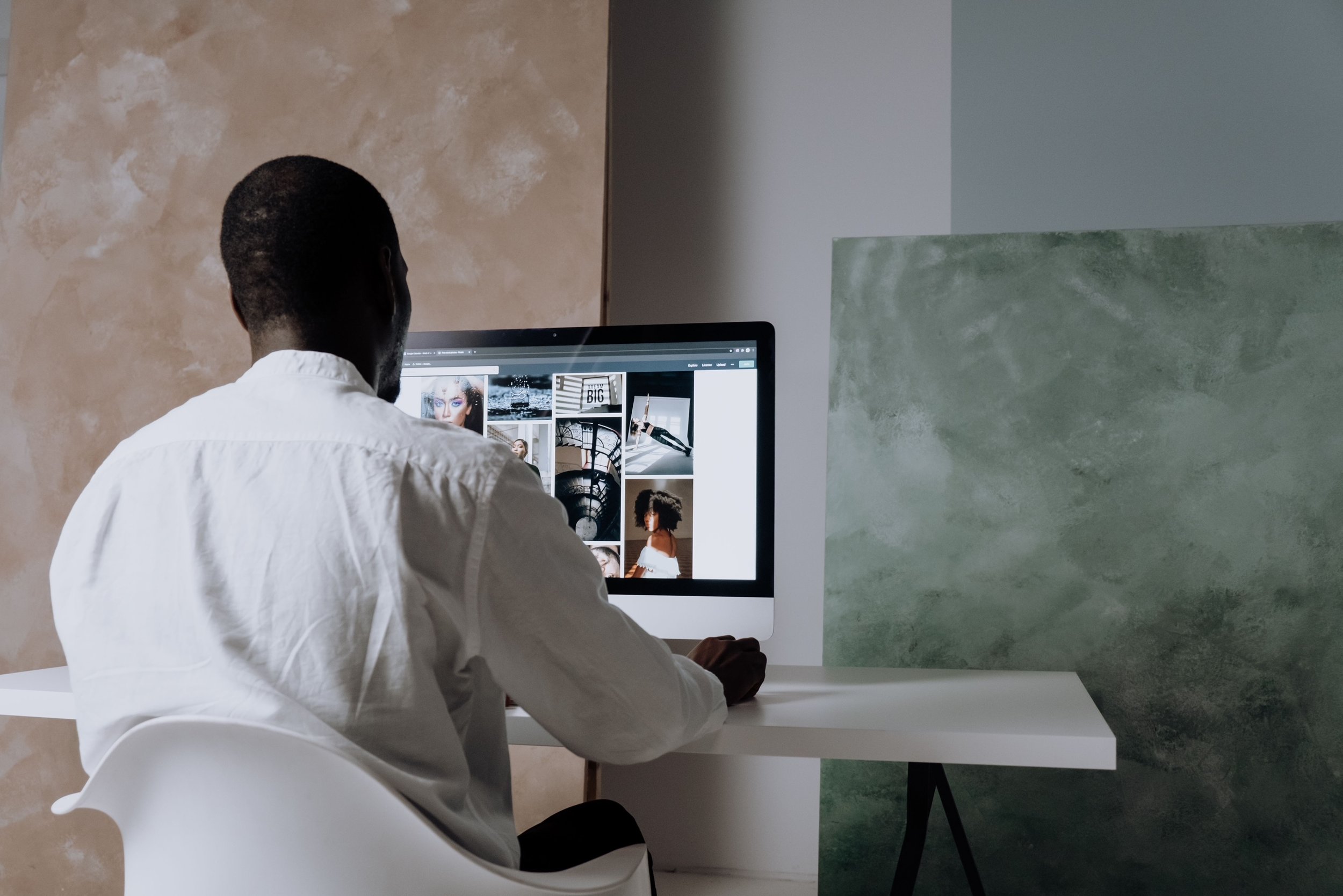 A person with dark skin and curly hair, wearing a white shirt, is sitting at a white desk in front of a computer monitor. The monitor displays a grid of images, including a portrait of a woman with makeup, architectural elements, and a woman with natural hair. The background features a beige textured wall and a green textured wall.