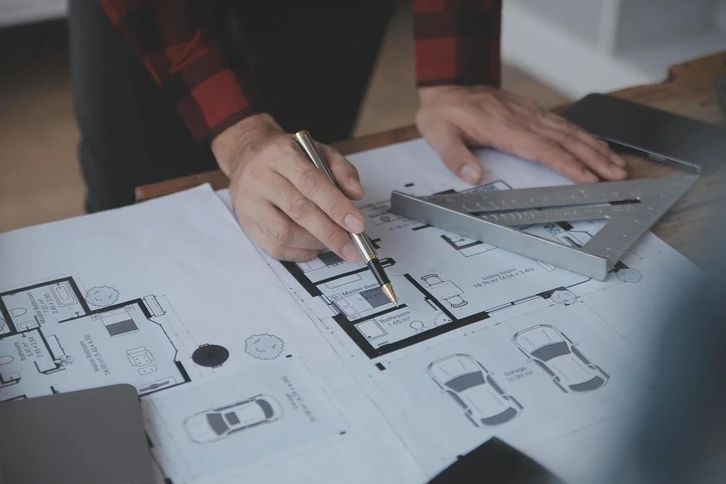 A person working on a house floor plan using a pen, metal ruler, and triangle on a wooden table.