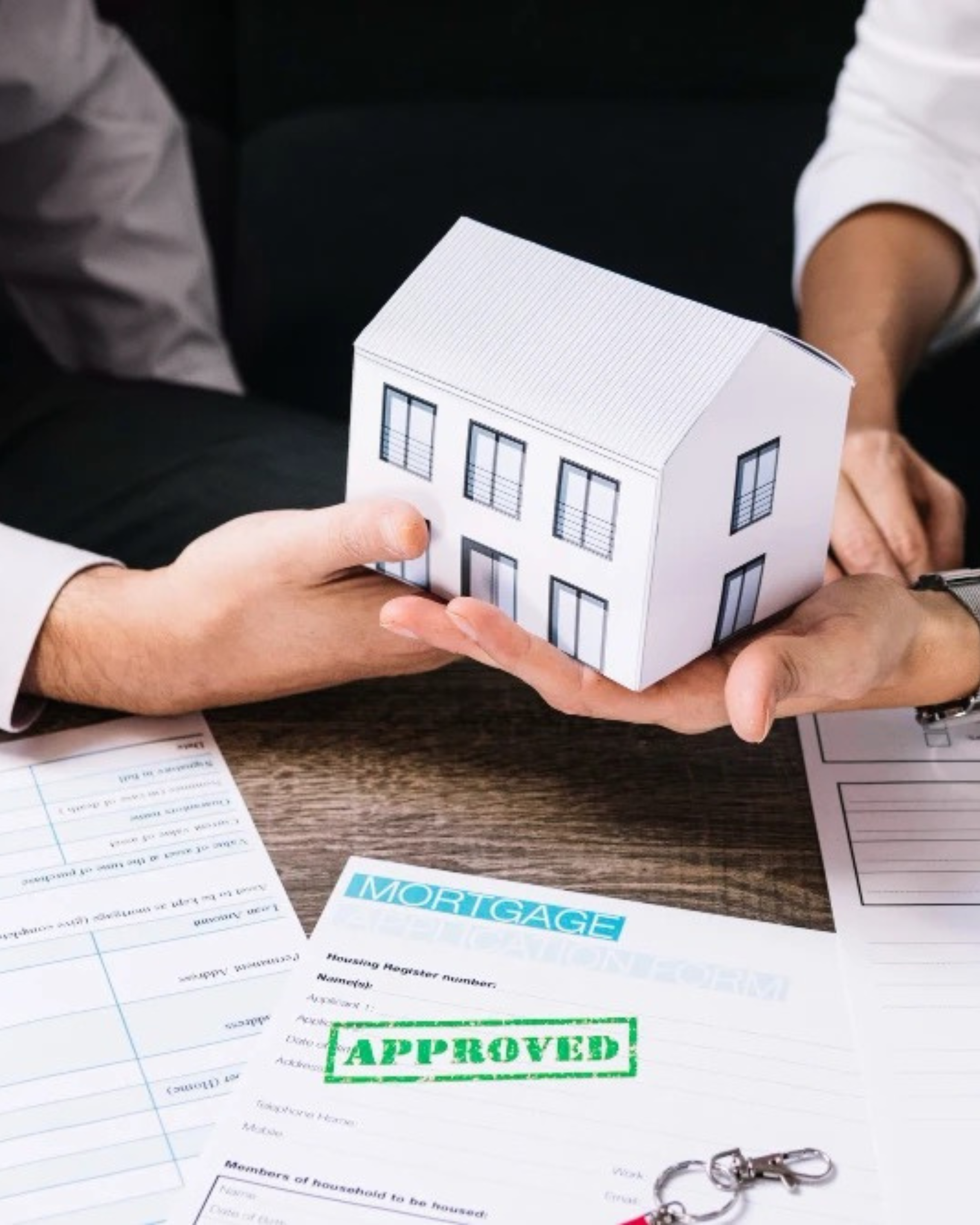 A group of people examining a small 3D paper model of a house. The model includes detailed windows and a roof. On the table, there are documents related to mortgages and approval status, including a green stamp with the word 'APPROVED' and a blue stamp with the word 'MORTGAGE'. A keychain with keys is also visible.