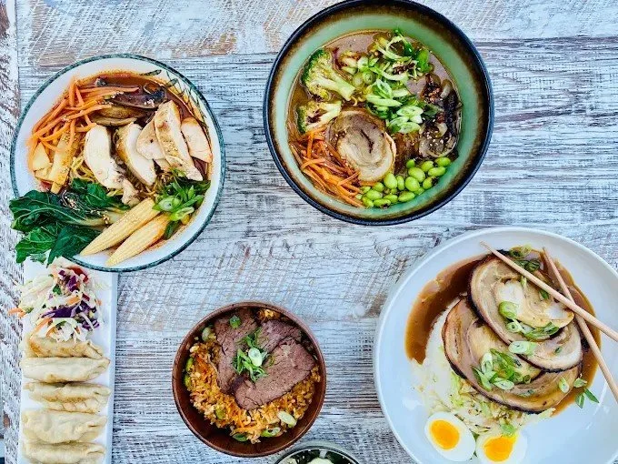 Assorted Asian dishes on a rustic wooden table, including bowls of ramen with sliced pork, vegetables, and noodles, a plate of dumplings, and a small bowl of minced beef topped with green onions.