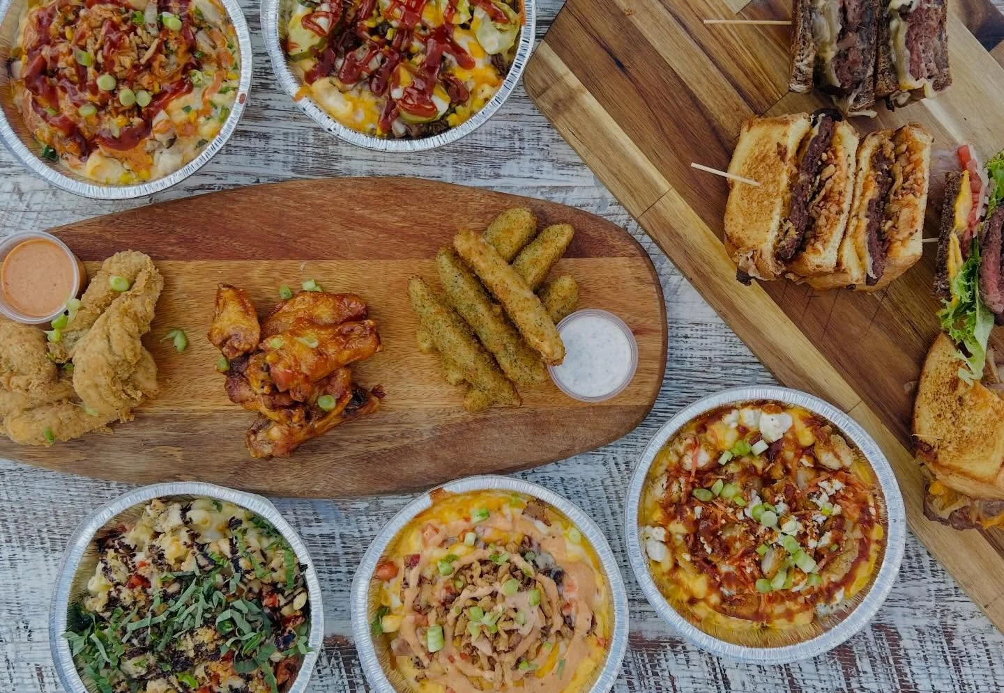 A variety of food including multiple small pizzas in metal trays, fried chicken, buffalo wing, fried breaded zucchini with dipping sauces, and large sandwiches on wooden serving boards.