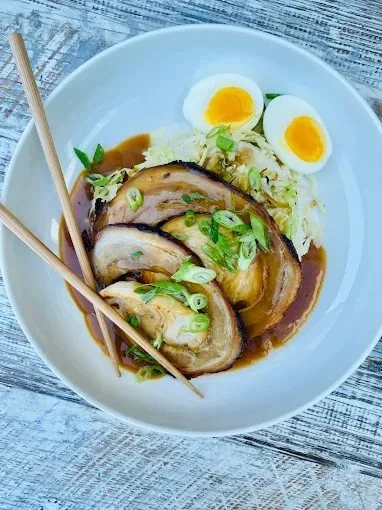 Bowl of ramen featuring sliced pork, halved soft-boiled eggs, chopped green onions, and broth, with wooden chopsticks resting on the side.