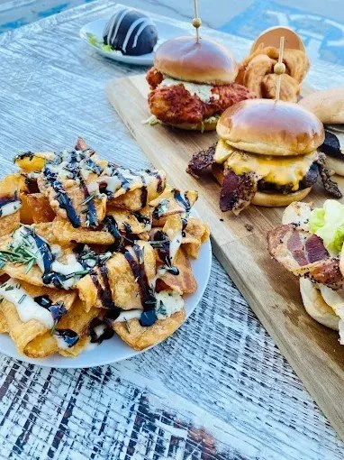 A plate of loaded fries topped with drizzled blue cheese and barbeque sauce, beside a wooden serving board with cheeseburgers, chicken sandwiches, and fried chicken with breaded chicken pieces, and a dessert on the table.