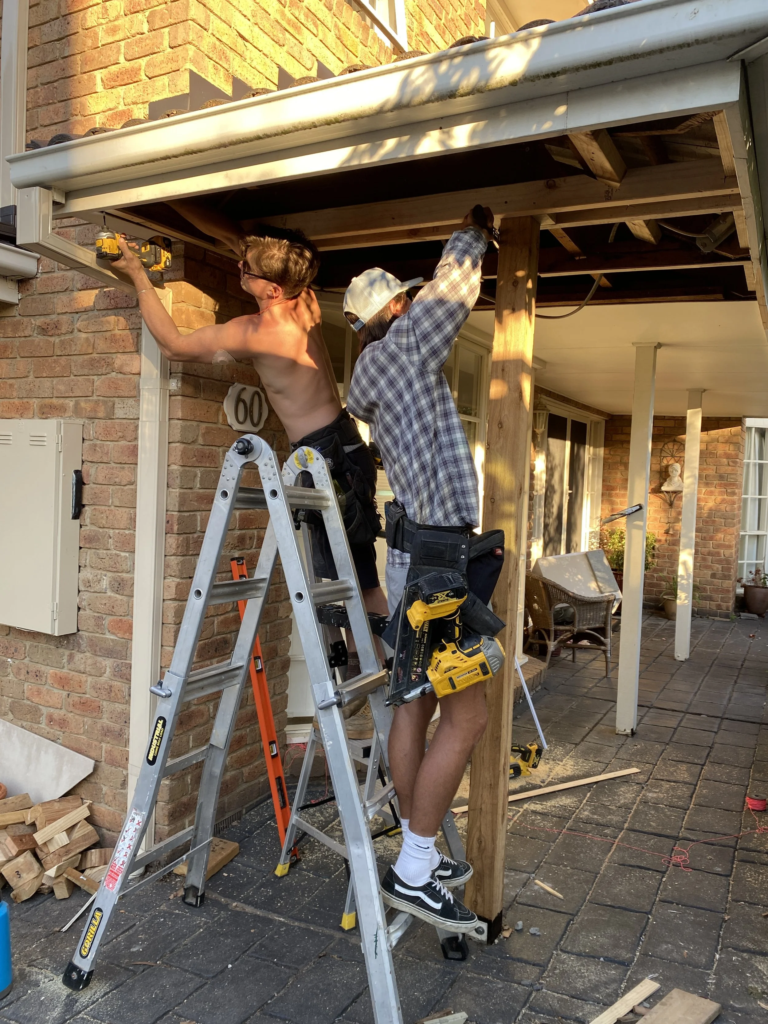 Two men working on building an outdoor structure. One man is shirtless, standing on a ladder, holding a power drill. The other man is wearing a plaid shirt, shorts, and a hat, working on the wooden frame. Construction tools and materials are scattered around them.