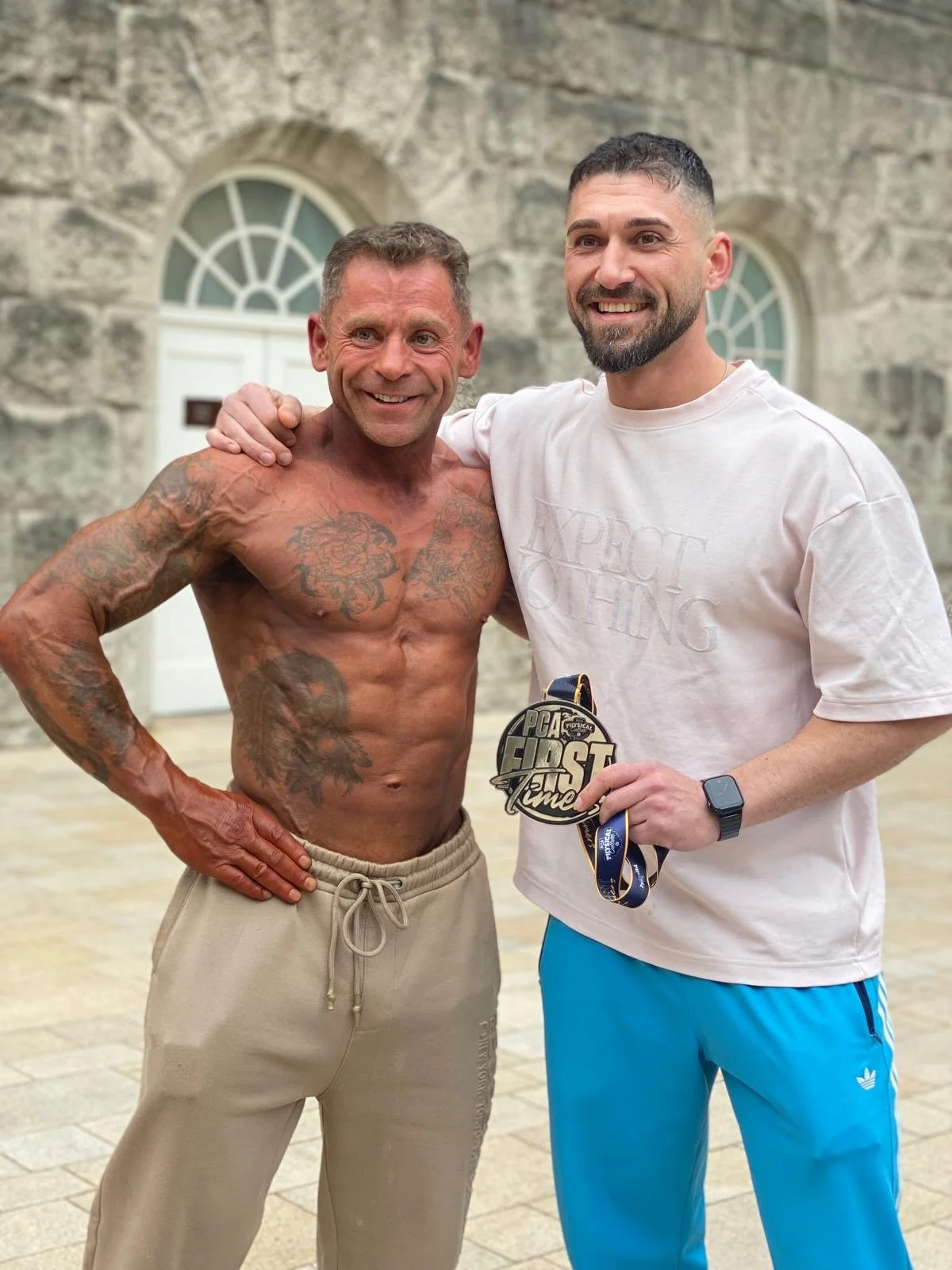 Two men standing together smiling outdoors, one shirtless showing tattoos, the other wearing a white t-shirt, blue pants, and holding a medal.