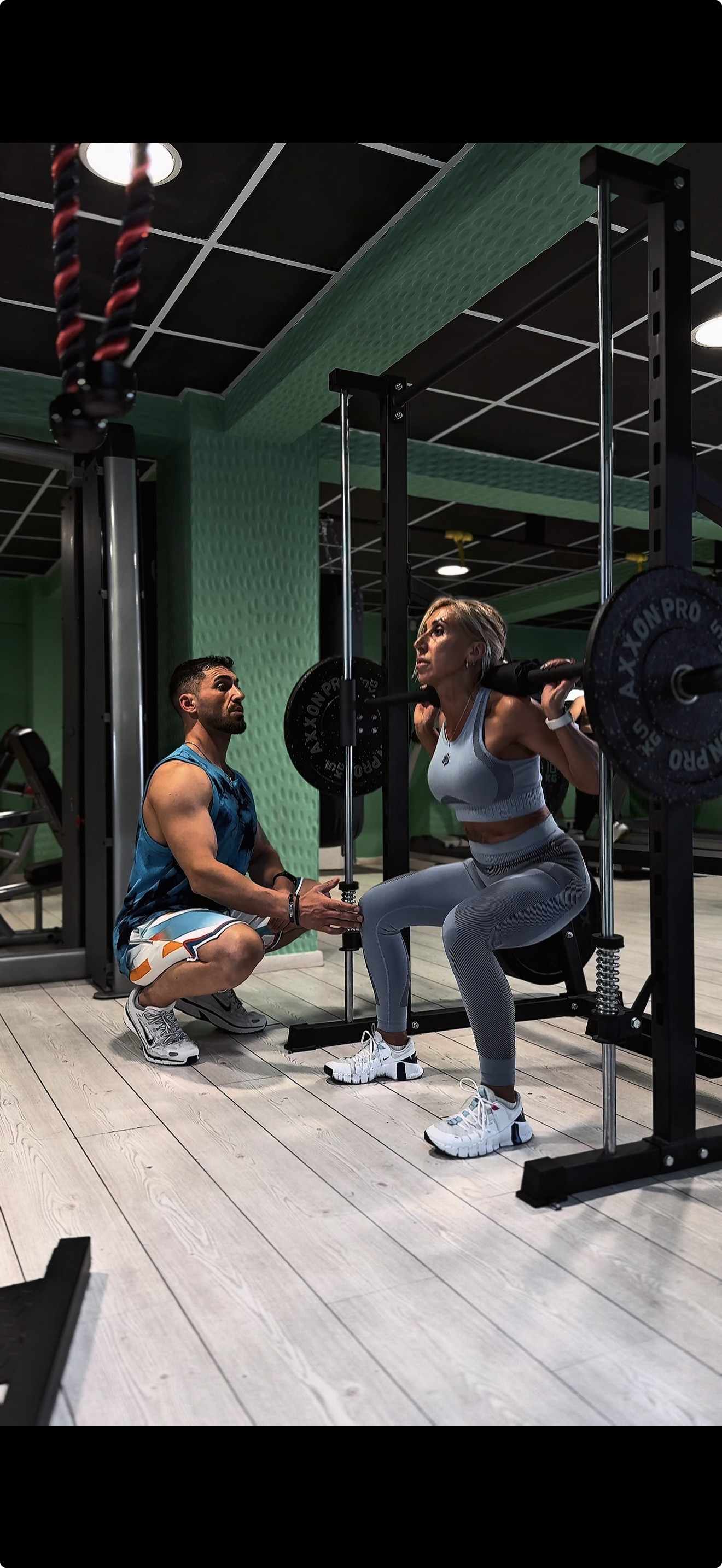 A woman performs a squat exercise with a barbell at a gym, supervised by a trainer kneeling beside her.