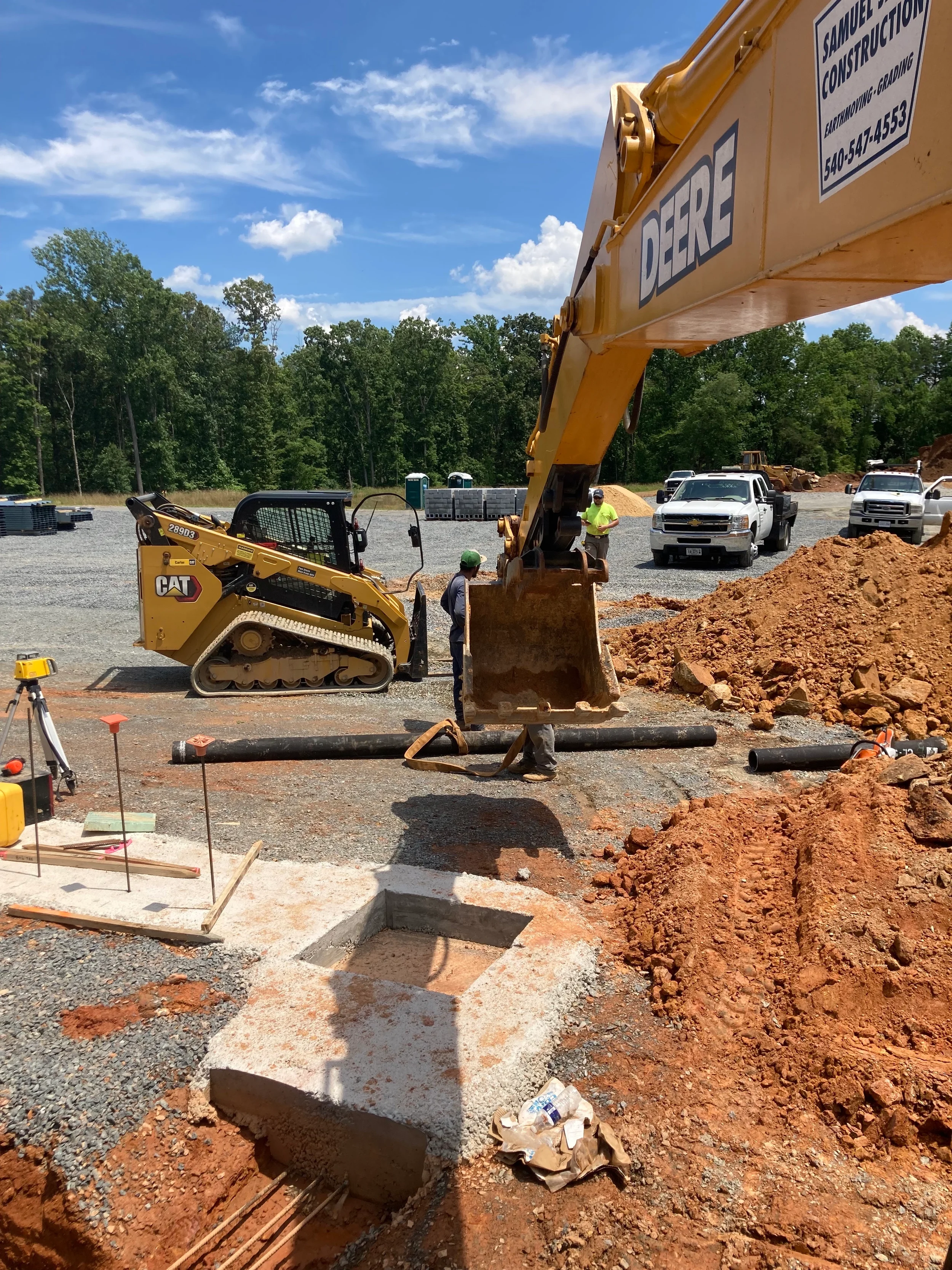 Commercial site pipe installation with excavator, skid steer, crew, stone base, trench area, and utility pipe staged for placement.