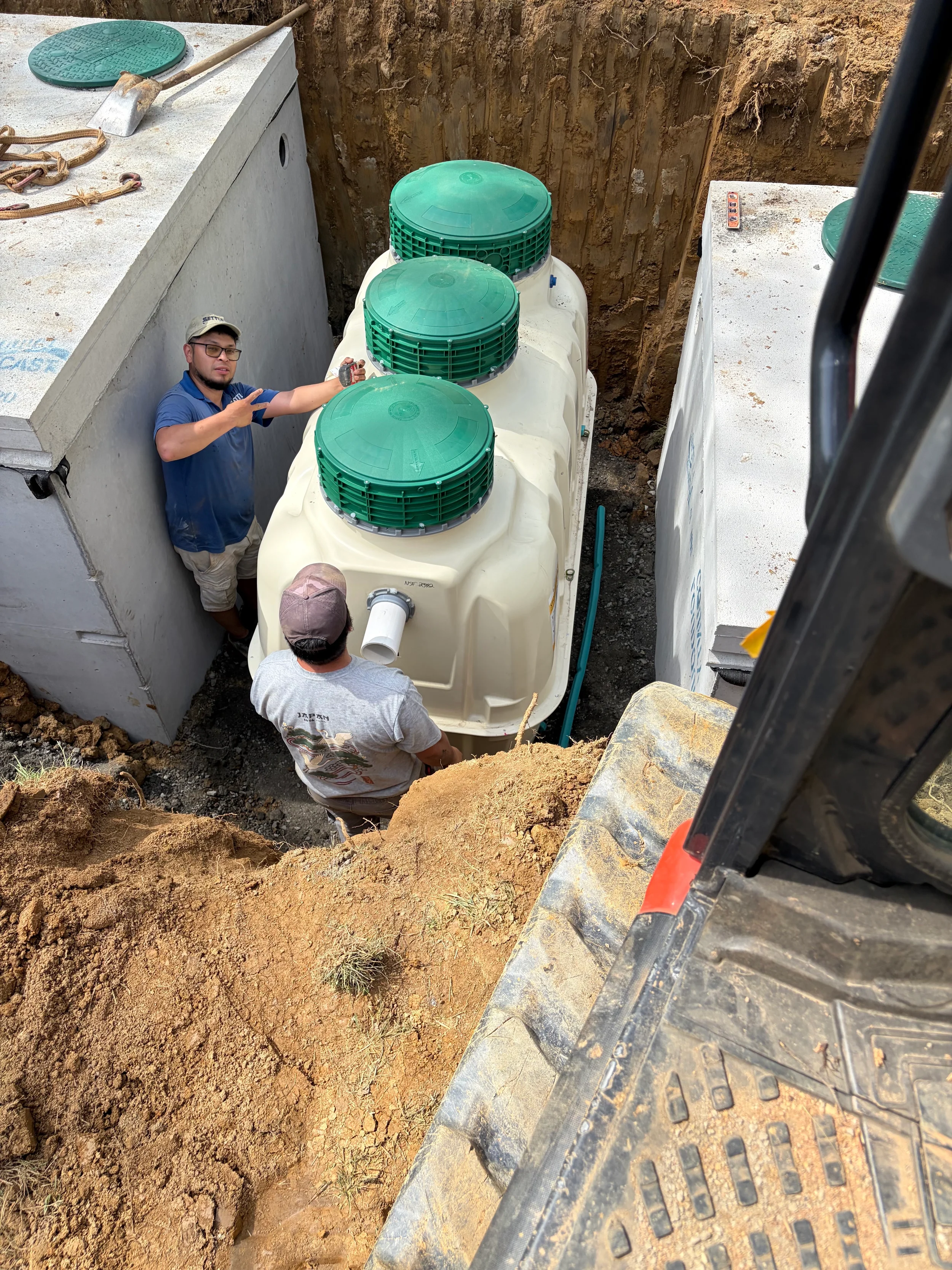 Workers position a poly septic tank between concrete structures during residential septic system installation and site excavation.