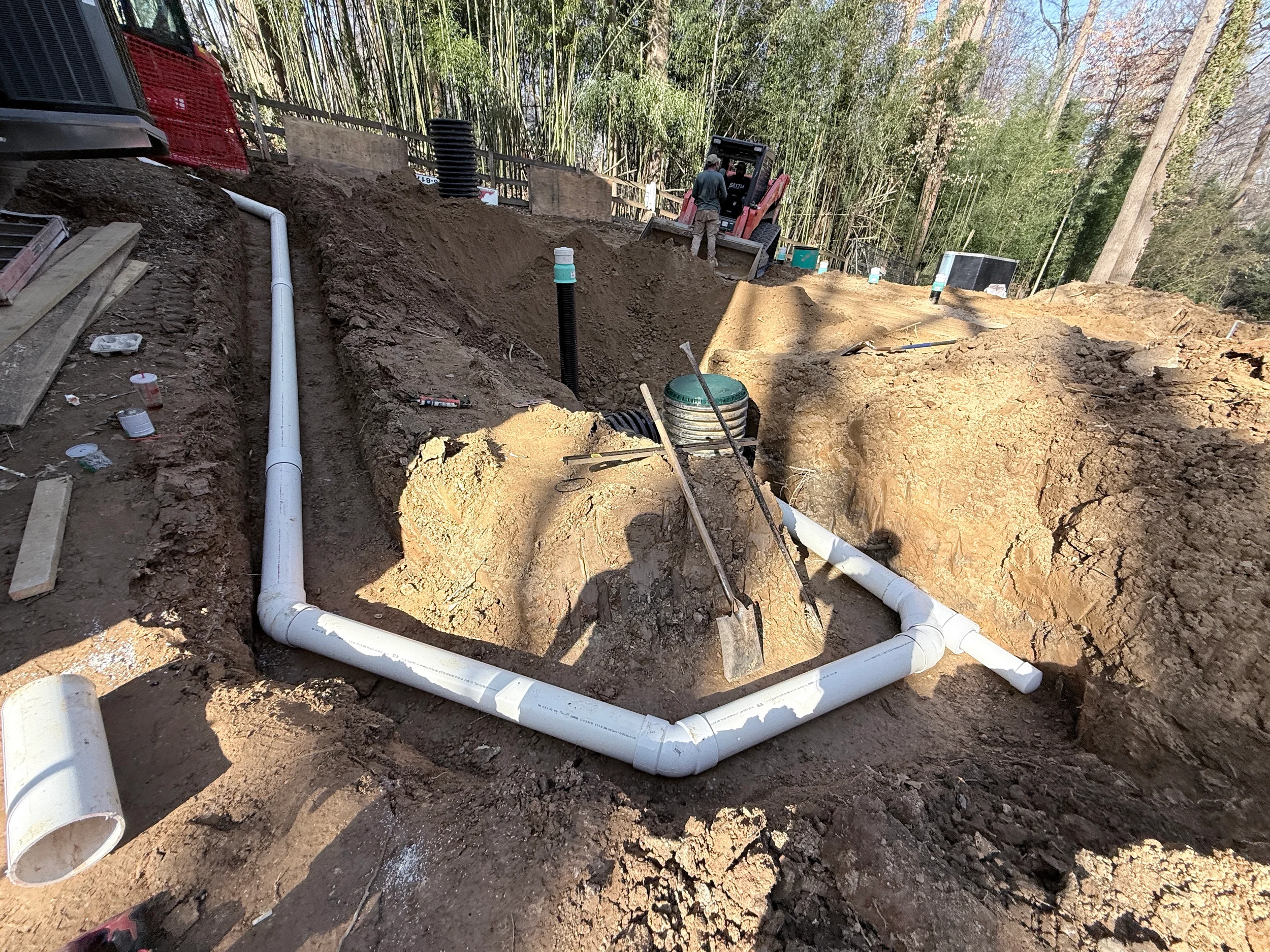 PVC stormwater pipe network with cleanout risers installed in an excavated trench beside a residential building.