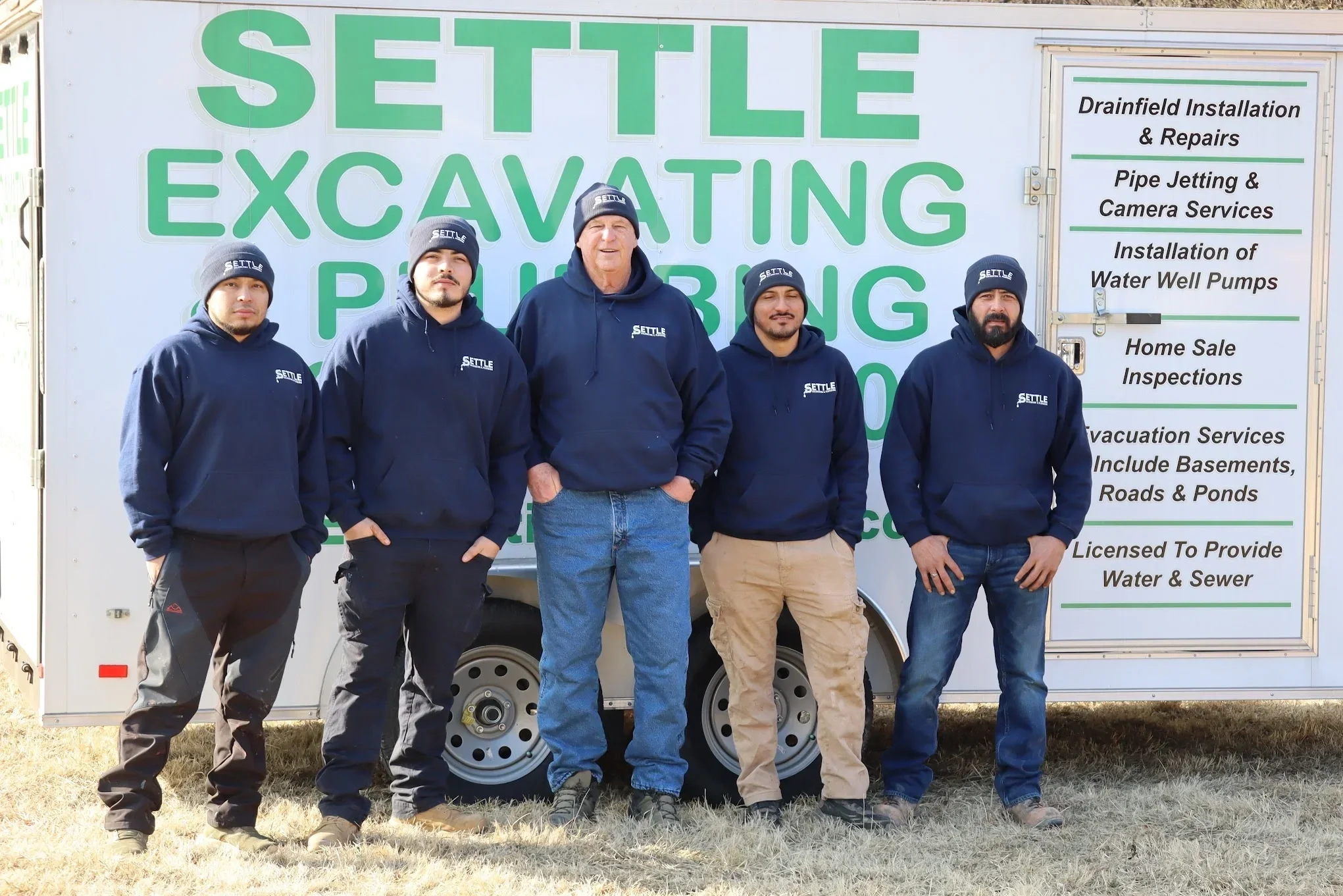 Settle Excavating & Plumbing crew standing in front of the company trailer wearing branded hoodies and winter hats.