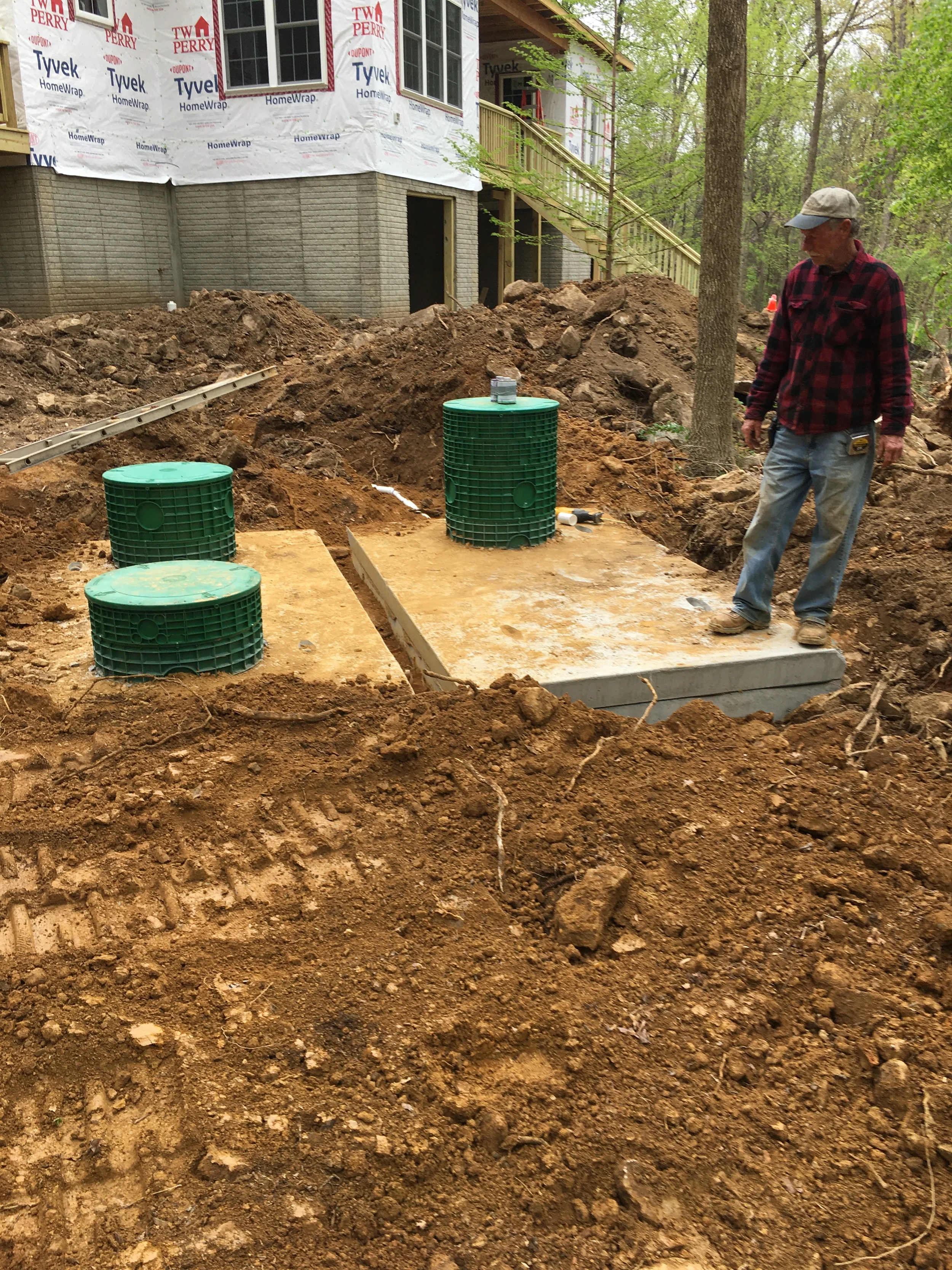 Septic tank riser installation at a new home site with concrete tank lids, green access risers, excavation, and rough site grading.