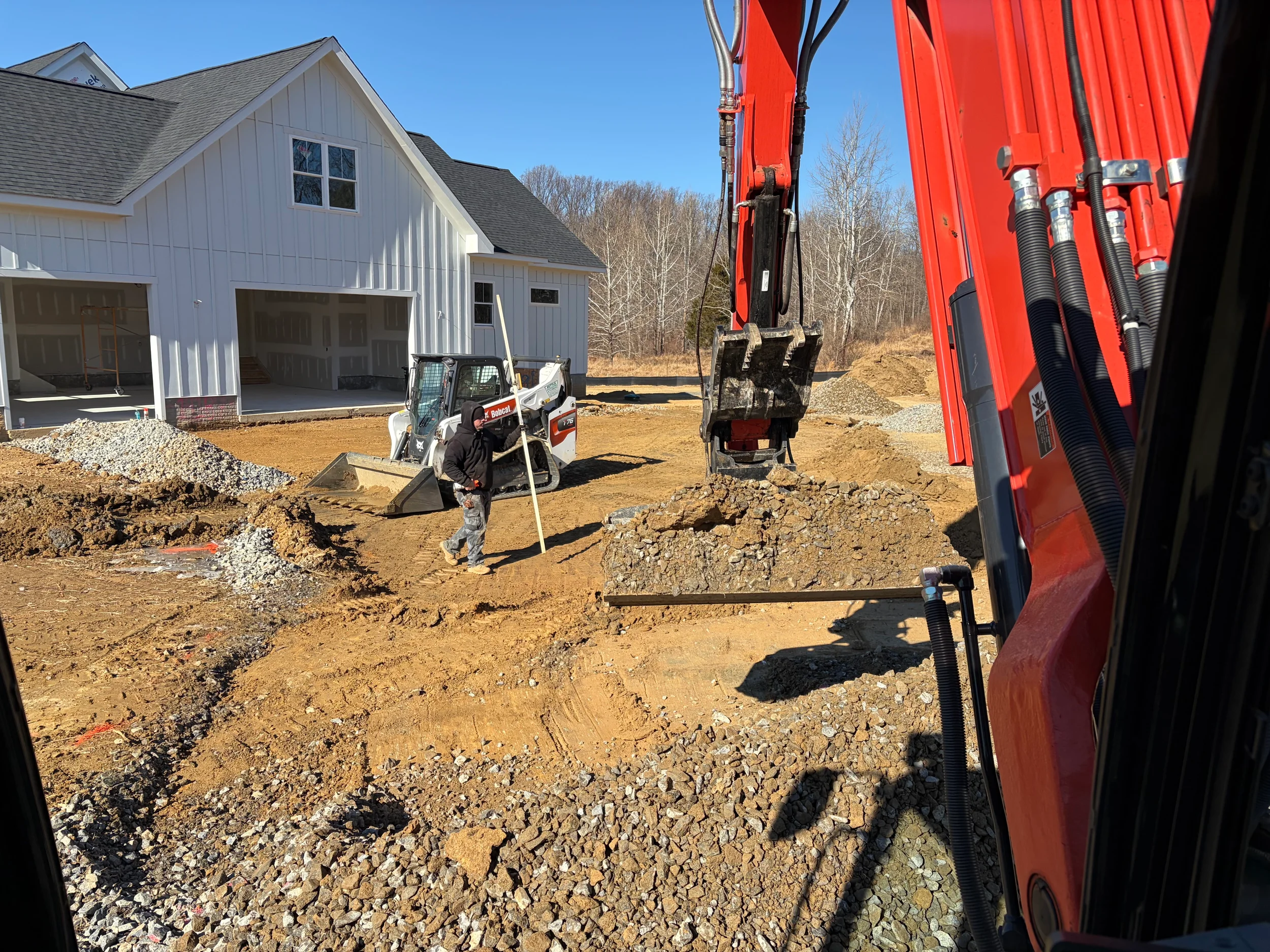 Excavator and Bobcat skid steer placing stone and shaping subgrade around a new farmhouse-style home under construction in Northern Virginia.