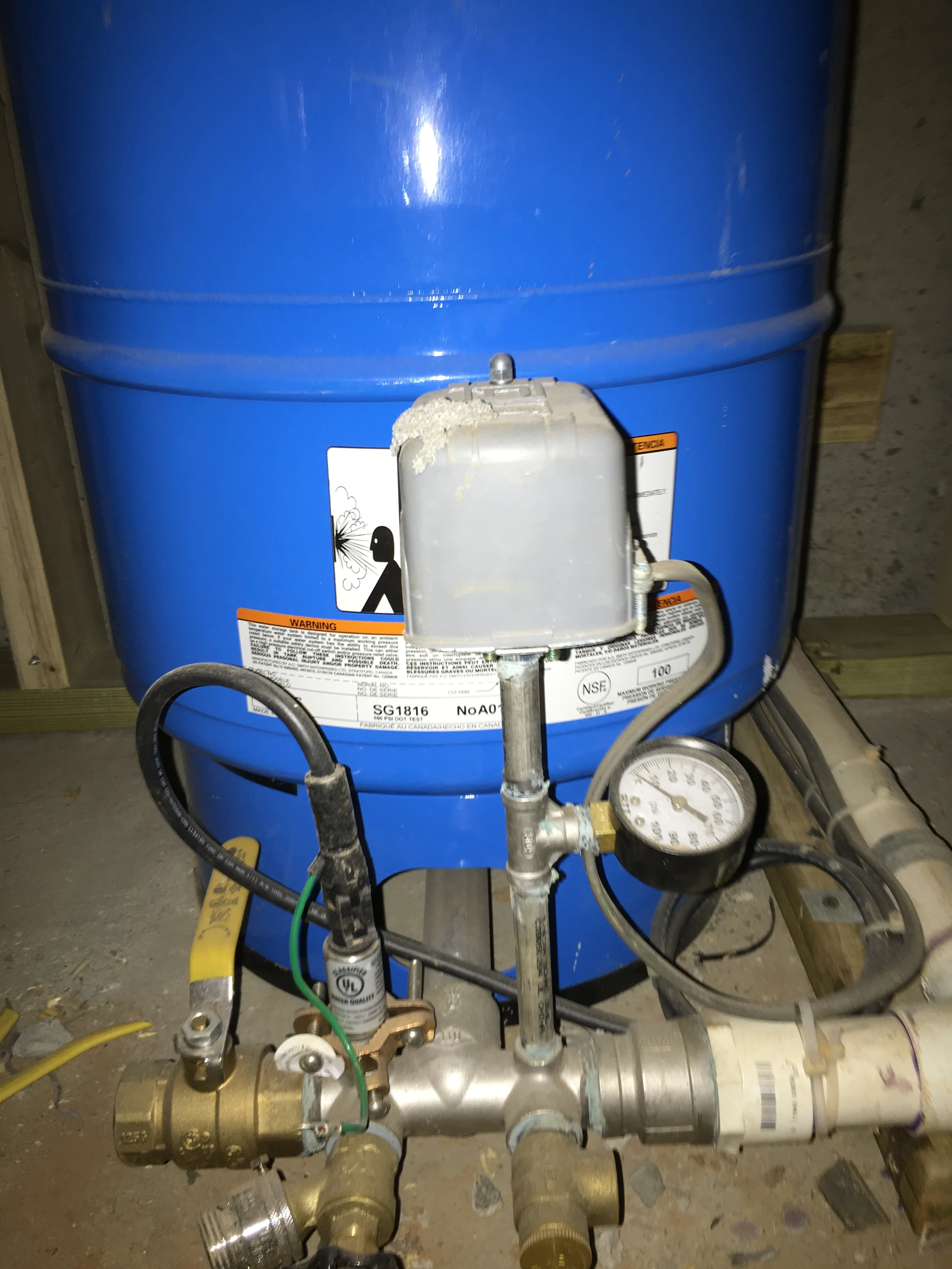 Closeup of a blue well pressure tank with pressure switch, pressure gauge, relief valve, and incoming water service piping.