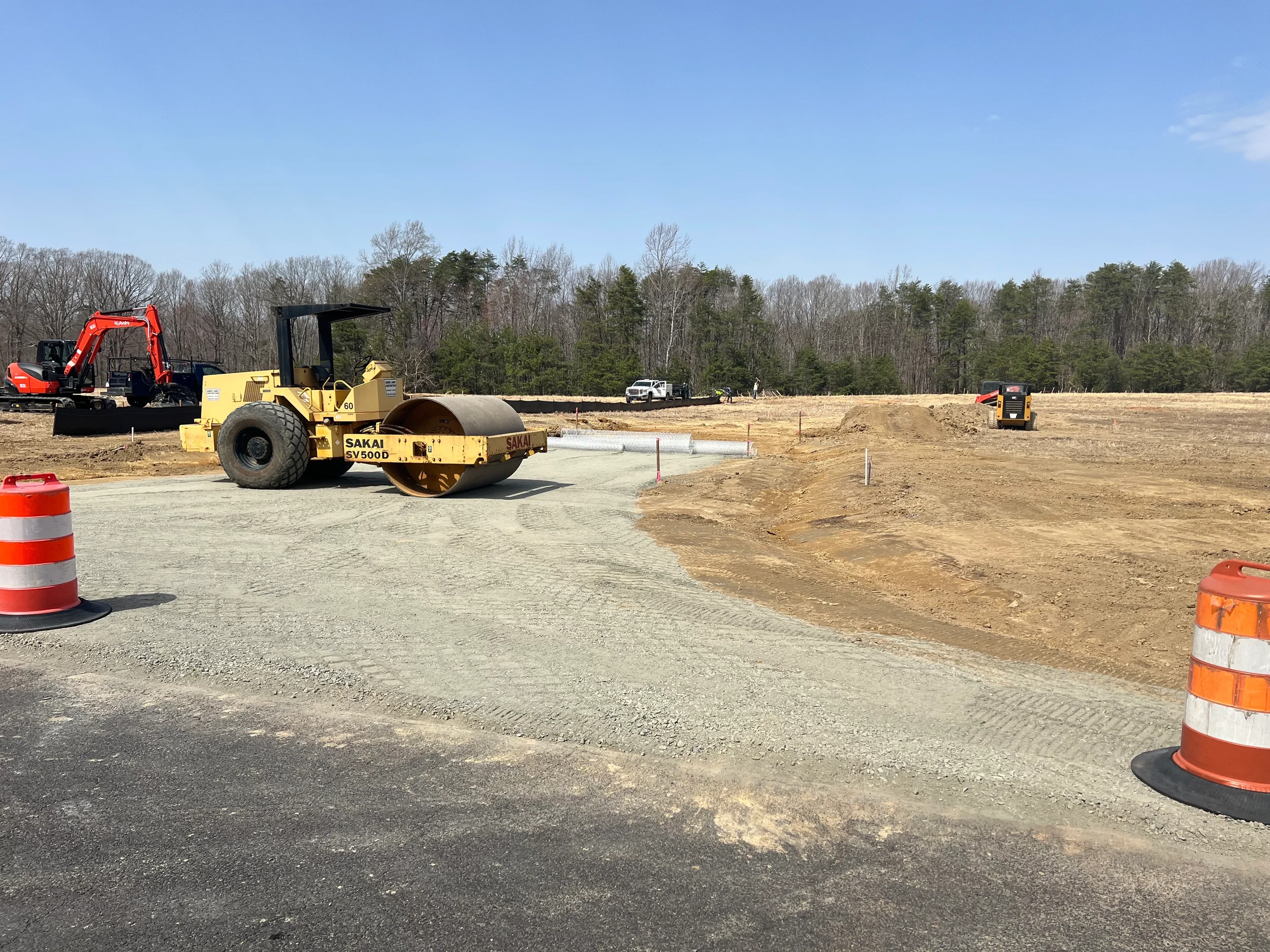 Sakai smooth-drum roller compacting a gravel construction entrance and access road, with excavators and silt fence on a Virginia site development project.