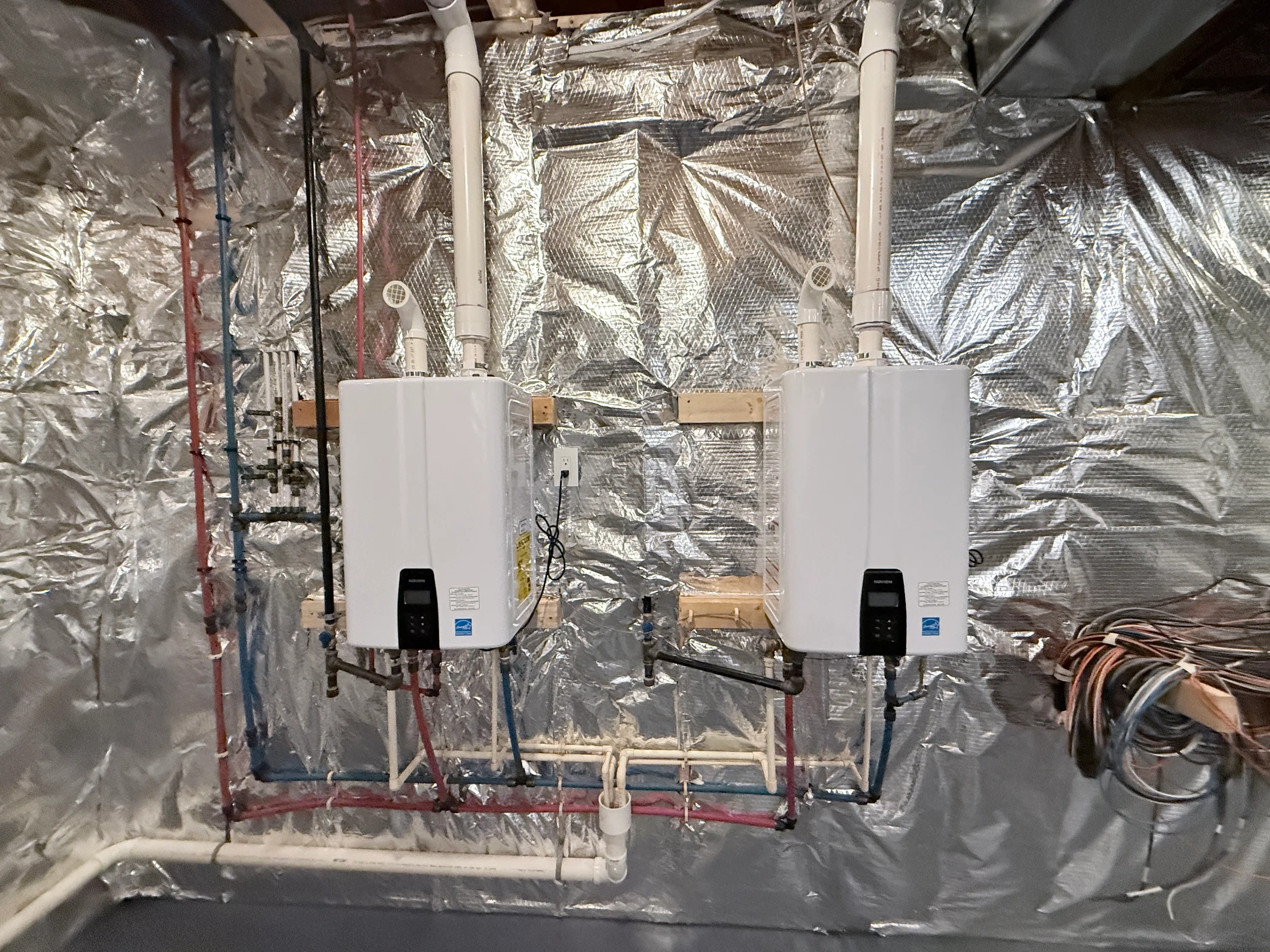 Two wall-mounted tankless water heaters plumbed with a red and blue PEX manifold and venting in an encapsulated crawlspace, a residential plumbing installation.