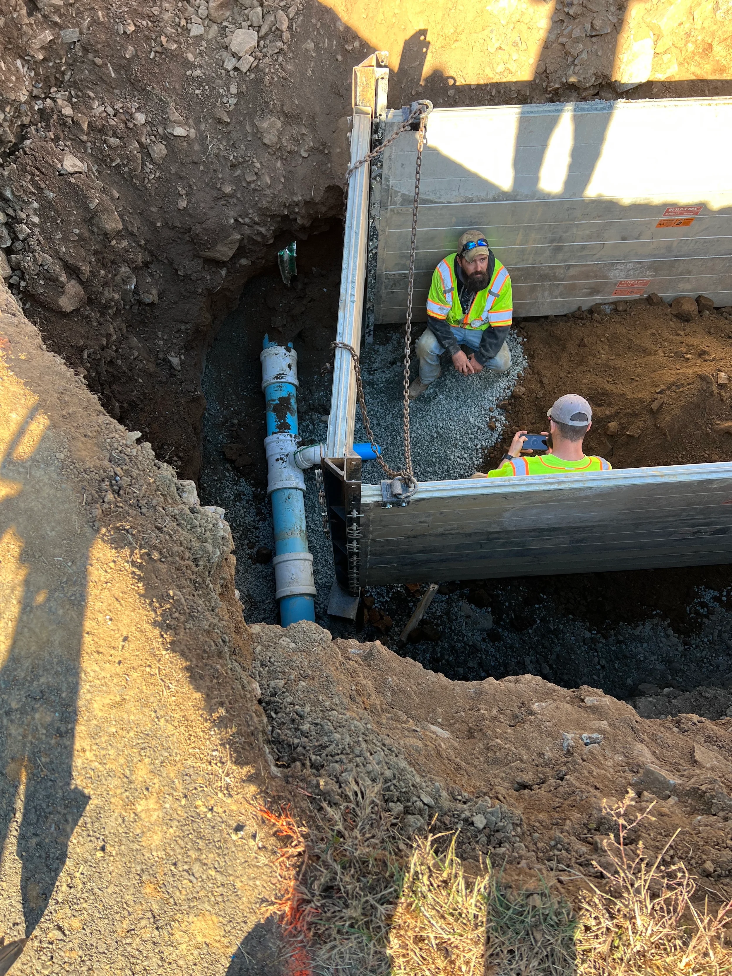 Water main repair with pipe bedding, trench shoring, utility crew documentation, gravel base, and exposed water pipe in excavation.
