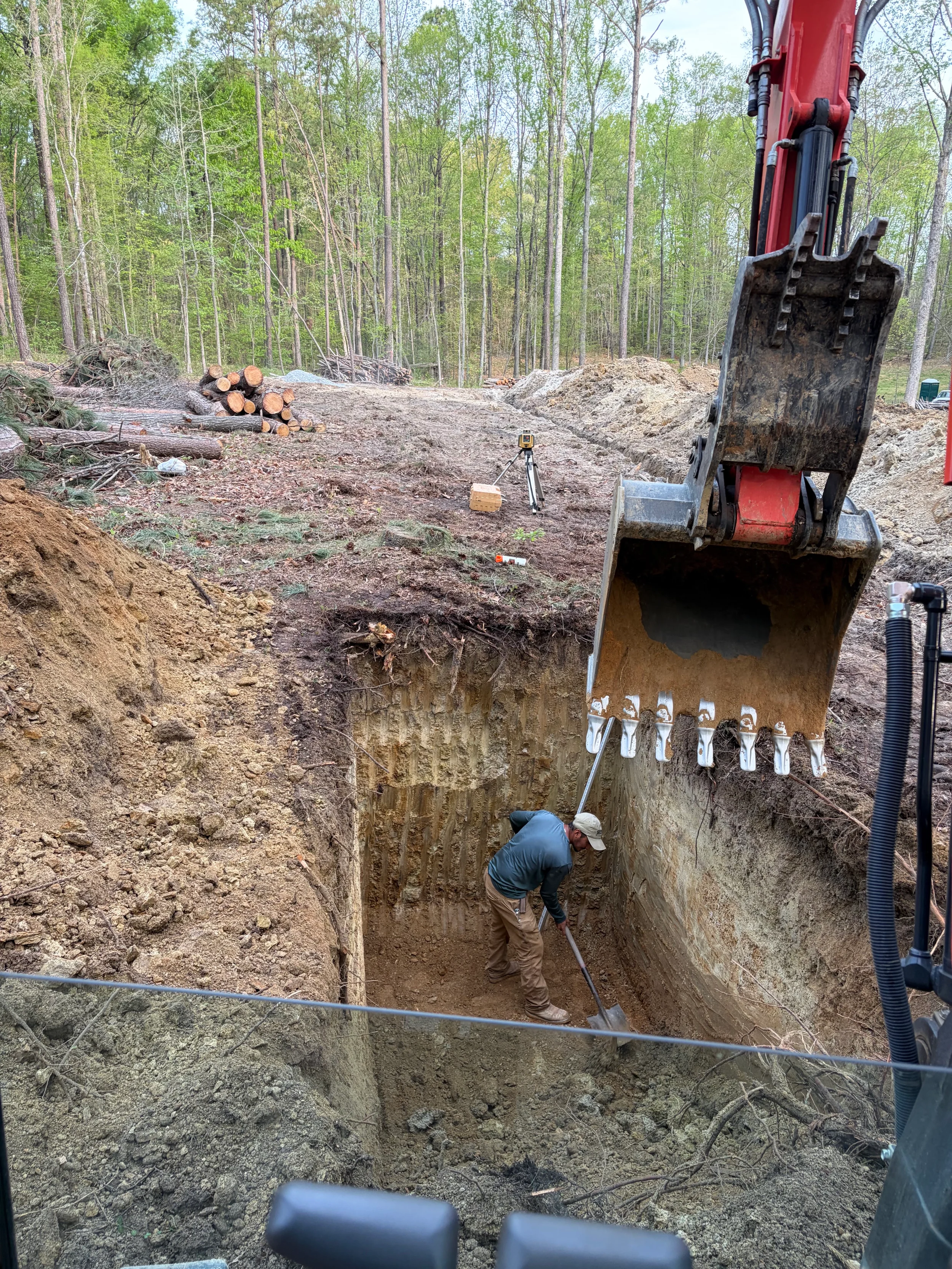 Deep excavation for septic tank installation with excavator bucket and worker preparing subgrade for residential system placement.