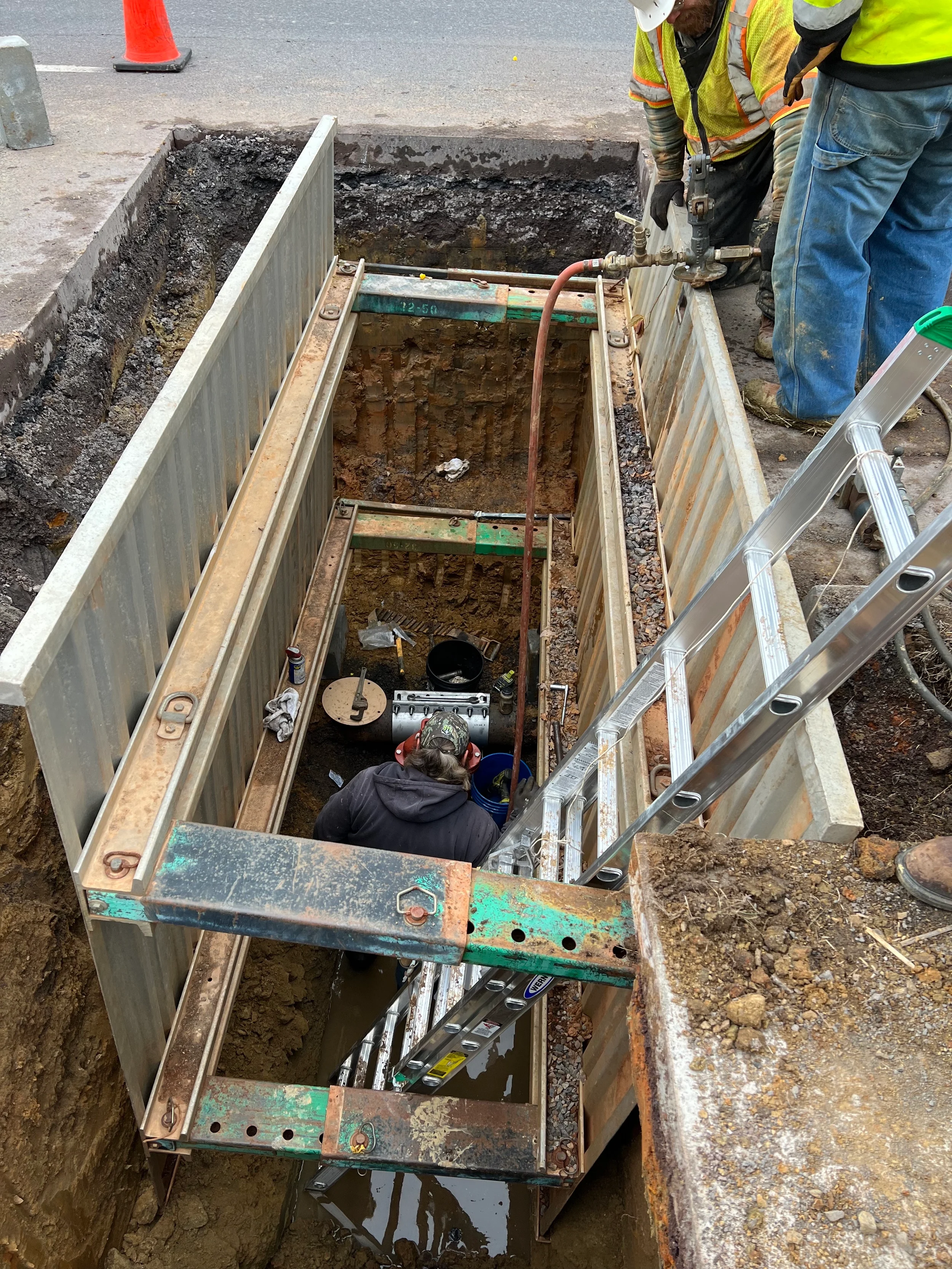 Water main tapping sleeve installation with crew working in a shored trench, valve connection, ladder access, and utility repair equipment.