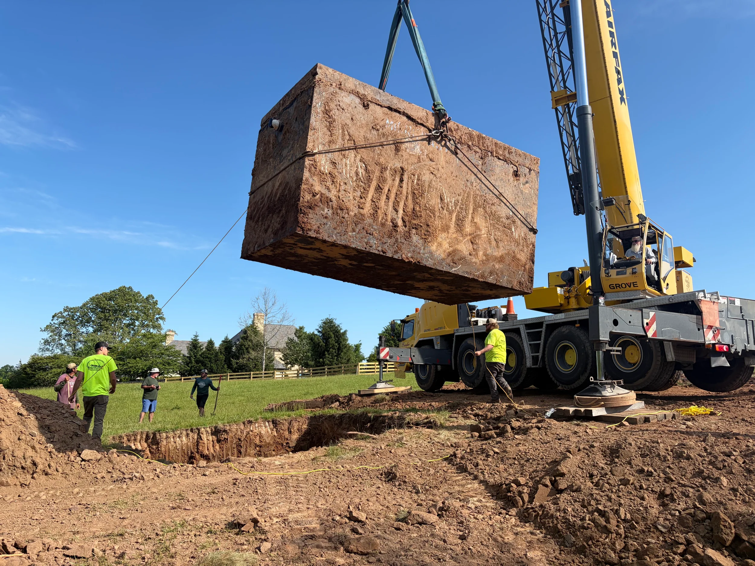 Crane placing a precast septic tank over an excavated area for residential septic installation with crew and equipment on site.