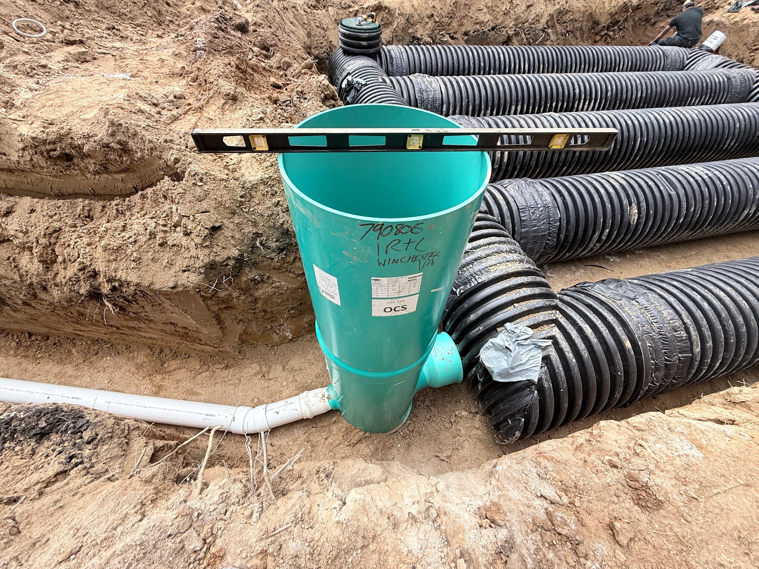 Green stormwater access riser with level, PVC connection, and HDPE detention pipe header inside an excavated underground system.