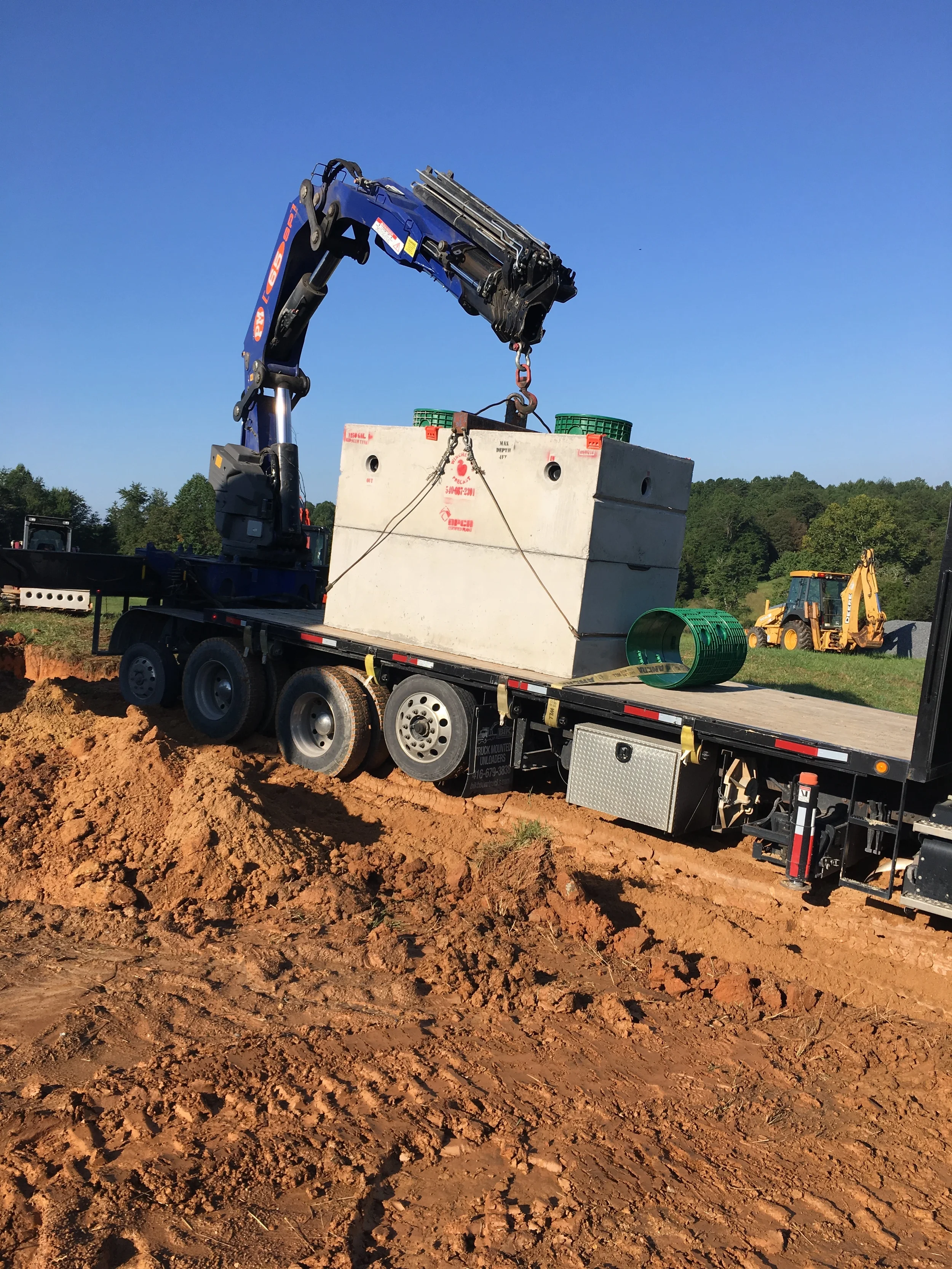 Truck mounted crane lifting a precast concrete septic tank with green risers off a flatbed trailer at a septic system install site in Northern Virginia.