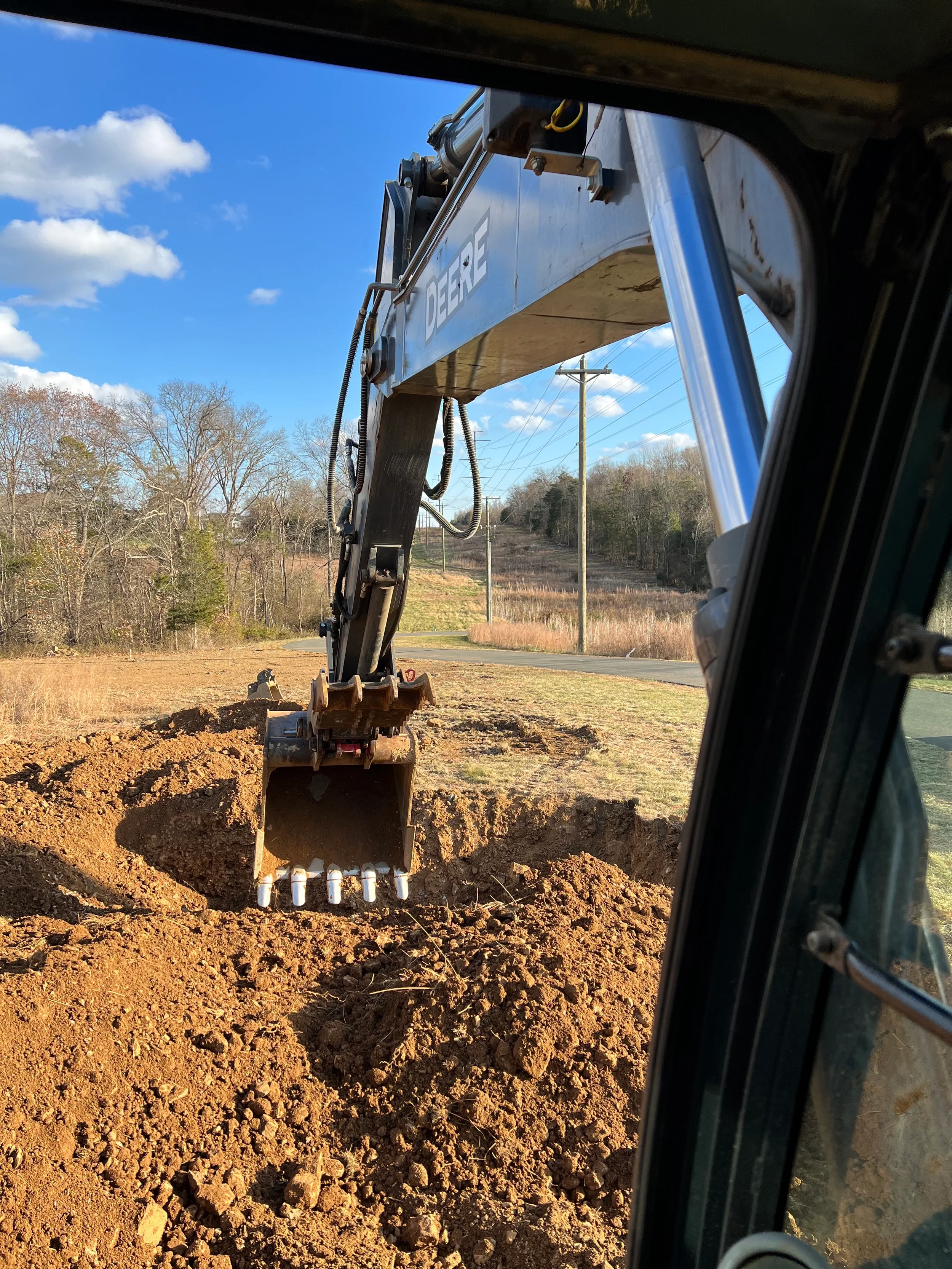 View from the cab of a John Deere excavator digging a utility trench through red Virginia clay along a residential driveway corridor on a site work project.